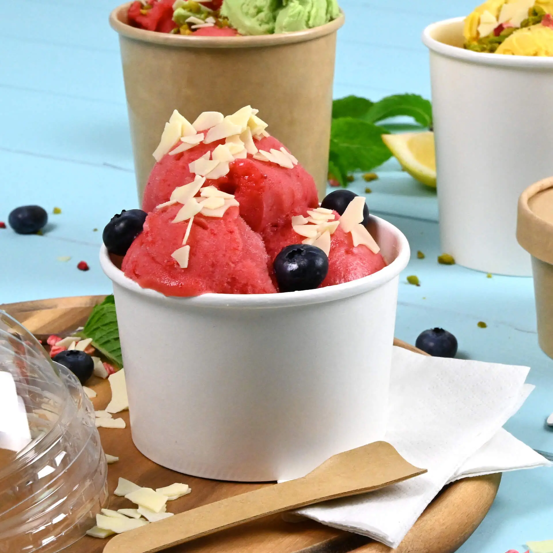 Biały kubek lodowy z powłoką PE wypełniony różowym sorbetem, udekorowany płatkami migdałowymi i borówkami, na drewnianej tacy.