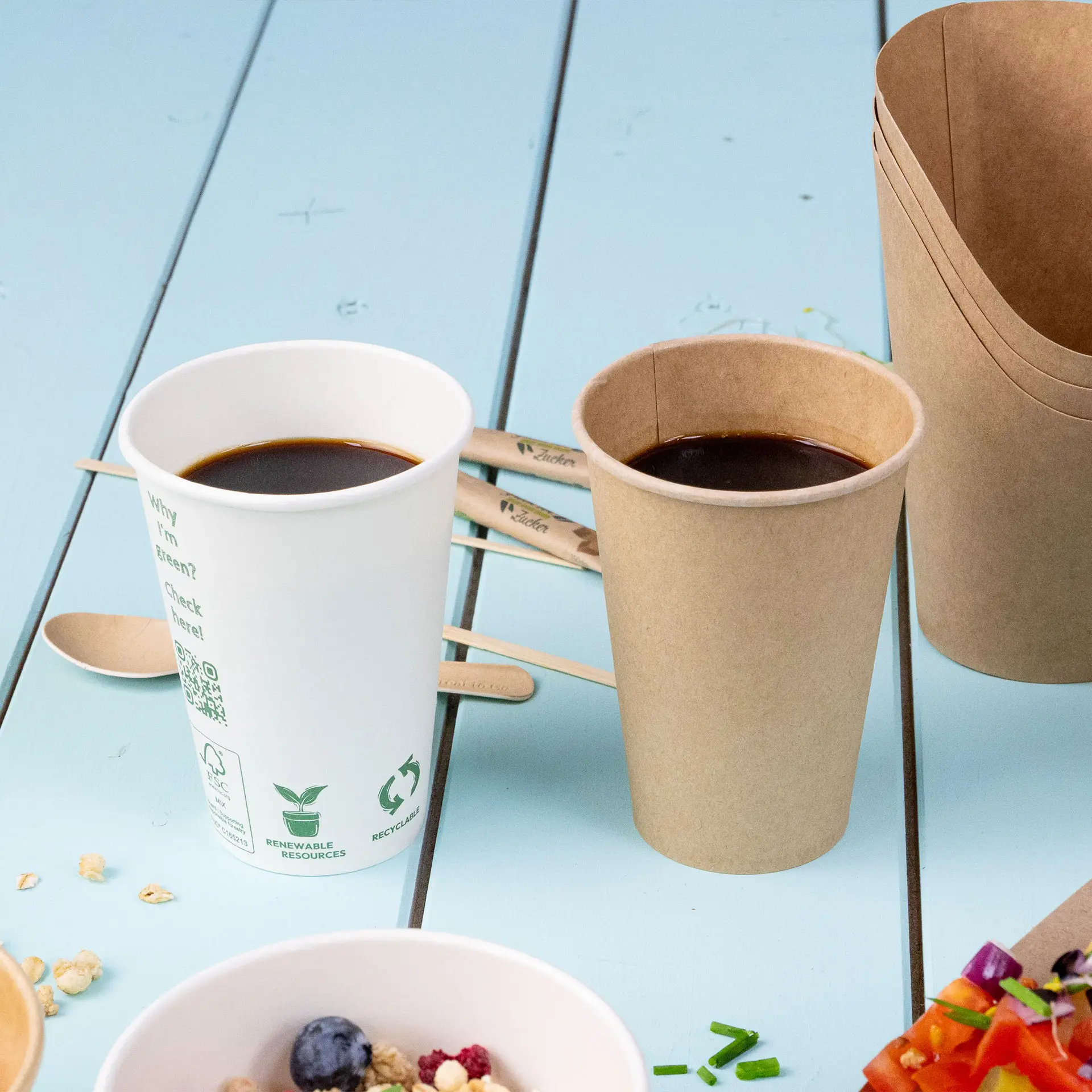 Jedna biała filiżanka z automatu wypełniona kawą i jedna brązowa filiżanka obok, z drewnianymi łyżkami w tle.