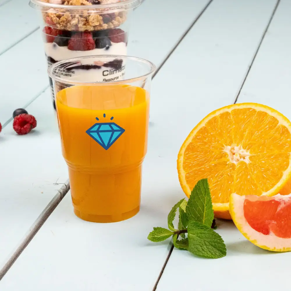 Przezroczysty kubek z sokiem pomarańczowym i niebieską grafiką diamentu, z plasterkiem pomarańczy i liśćmi mięty.