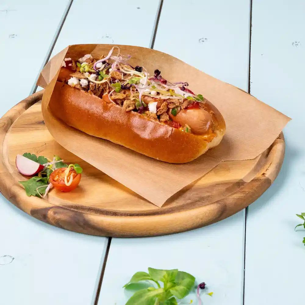 Hot dog w bułce z cebulą, ziołami i chrupiącymi dodatkami na drewnianej tacce z warzywami.