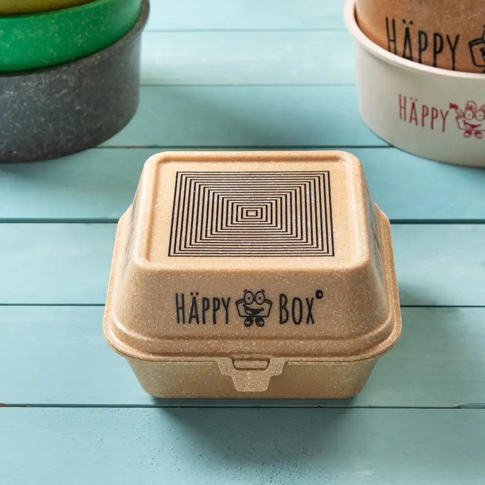 Wielokrotnie używany pojemnik w kwadratowym kształcie z designem pokrywki i logo "HAPPY BOX" na niebieskiej drewnianej powierzchni.