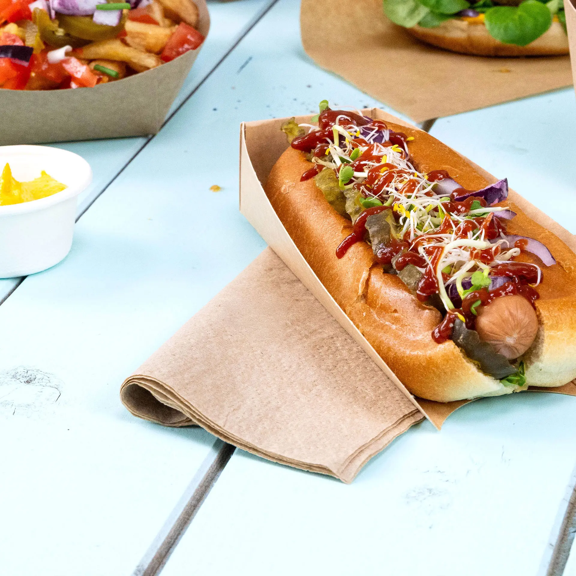 Hot dog w papierowym uchwycie, z ketchupem, musztardą, ogórkami i kiełkami, z serwetką obok na niebieskim stole.
