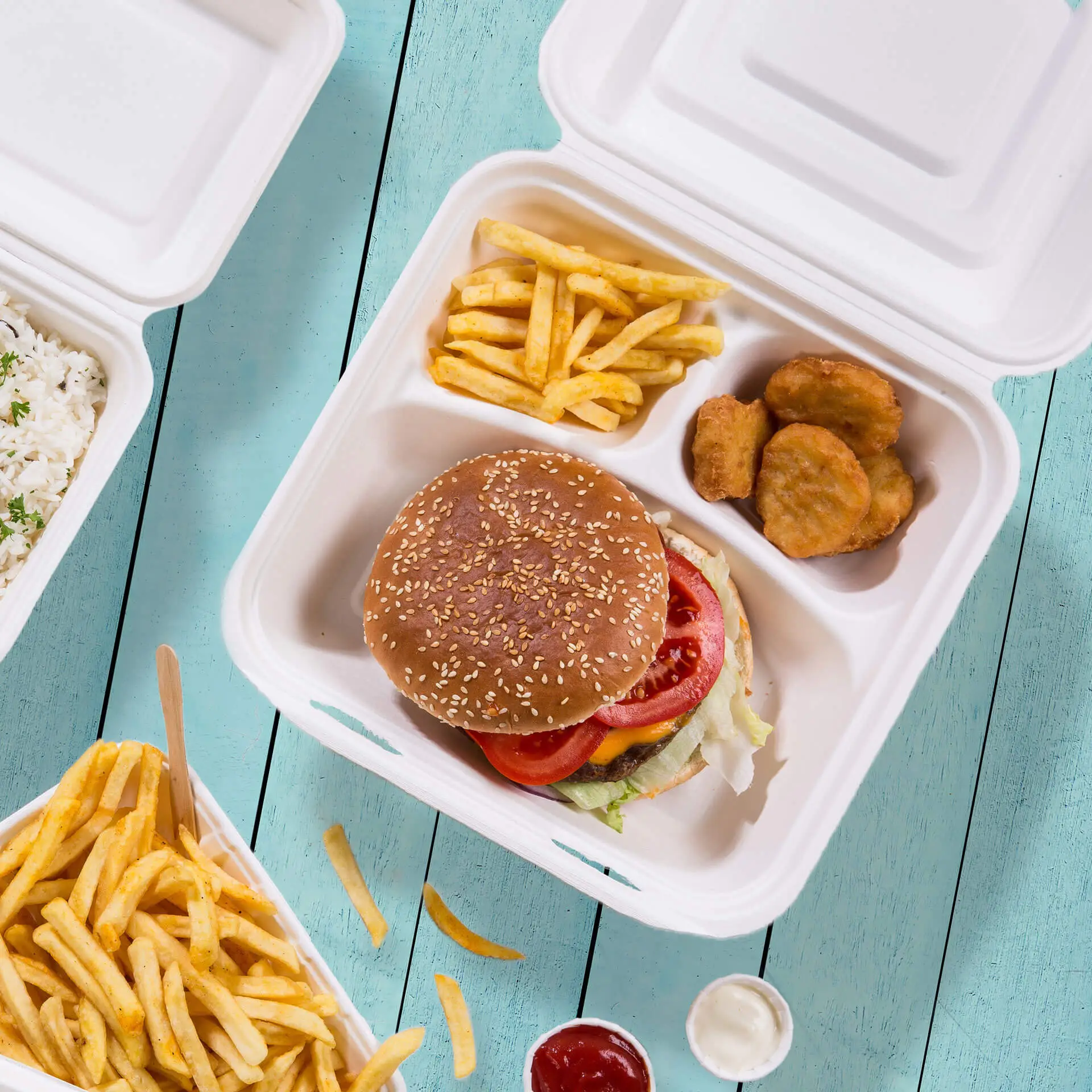 Biodegradowalna pudełko na menu z trzema przegrodami zawierające hamburgera z sezamem, frytki i nuggetsy kurczaka na niebieskim drewnianym stole.