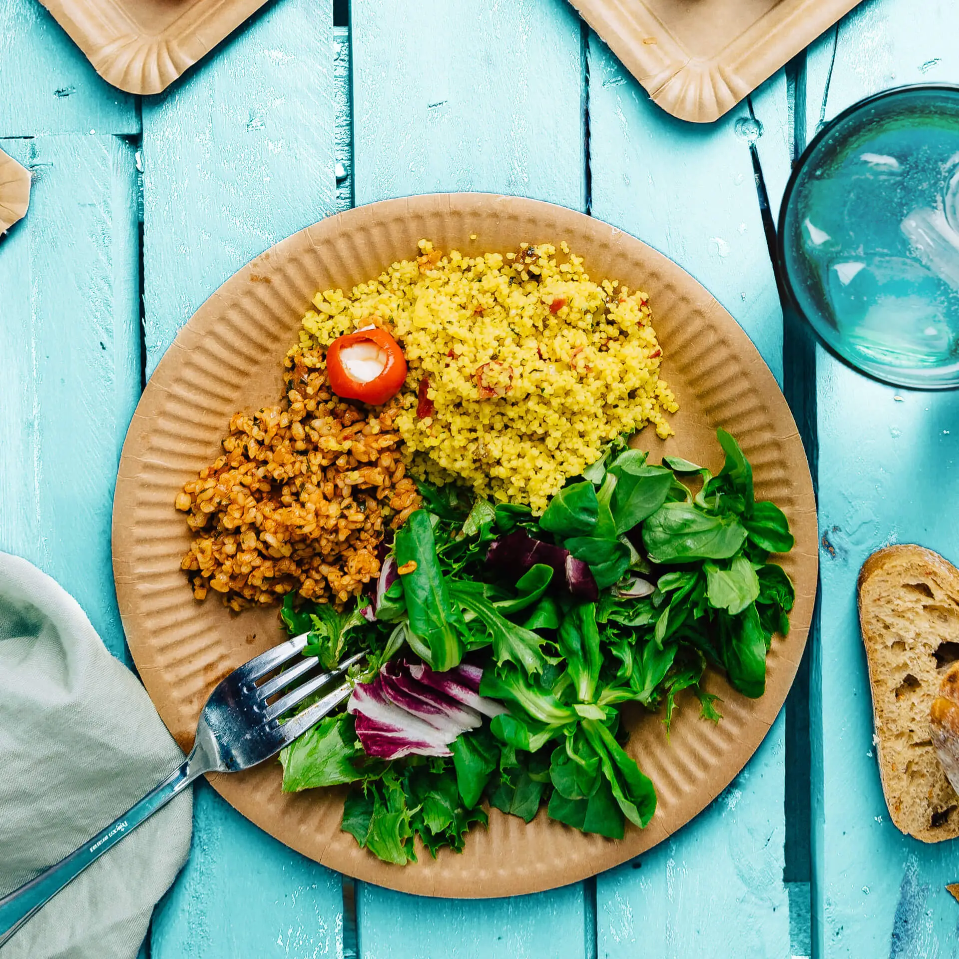 Okrągły brązowy talerz z tektury z żółtym kuskusem, brązowymi ziarnami i mieszanką zieleniny; widelec oparty na sałatce.