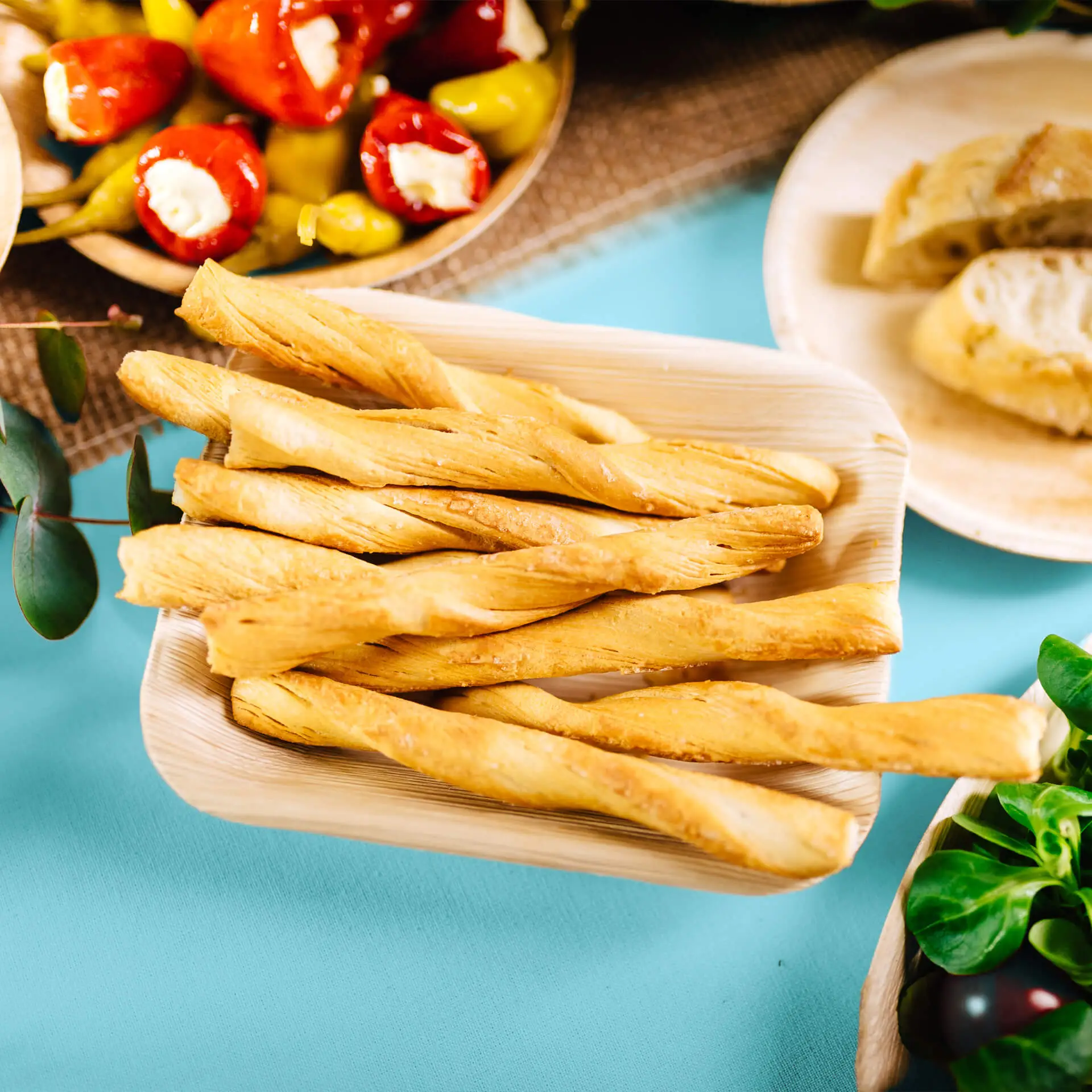 Talerz z liści palmowych trzymający kilka złotych skręconych paluszków chlebowych, z dodatkowymi jedzeniem rozmytym w tle.