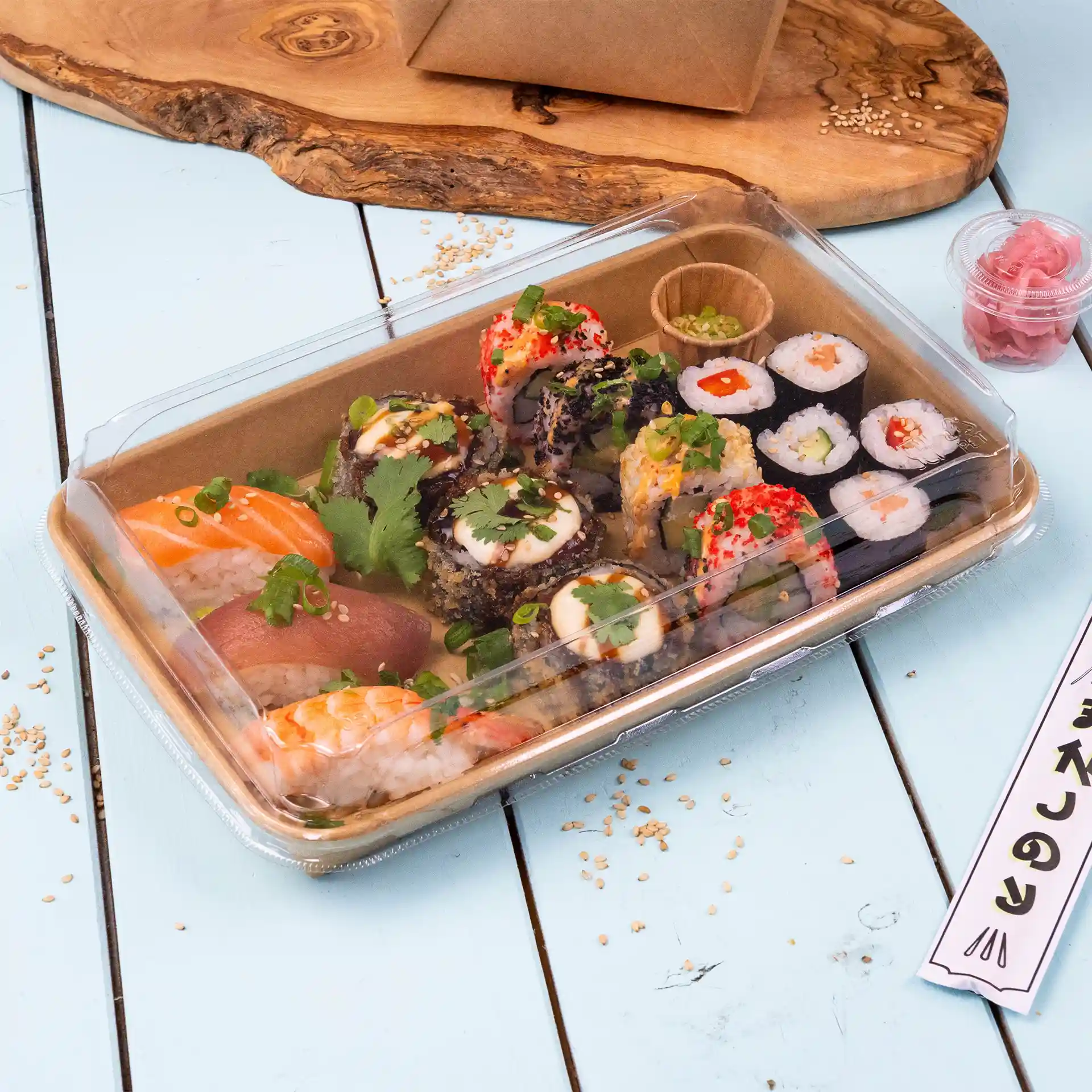 Przezroczysta pokrywka na pudełko sushi z różnymi rolkami sushi, w tym łososiem i tuńczykiem, udekorowanymi ziołami i podawanymi z marynowanym imbirem.