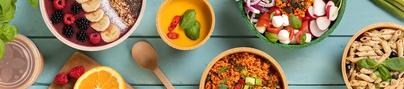 Kolorowe miski wypełnione różnymi potrawami, w tym owocami, makaronem, sałatką i zbożami, ułożone na drewnianym stole.