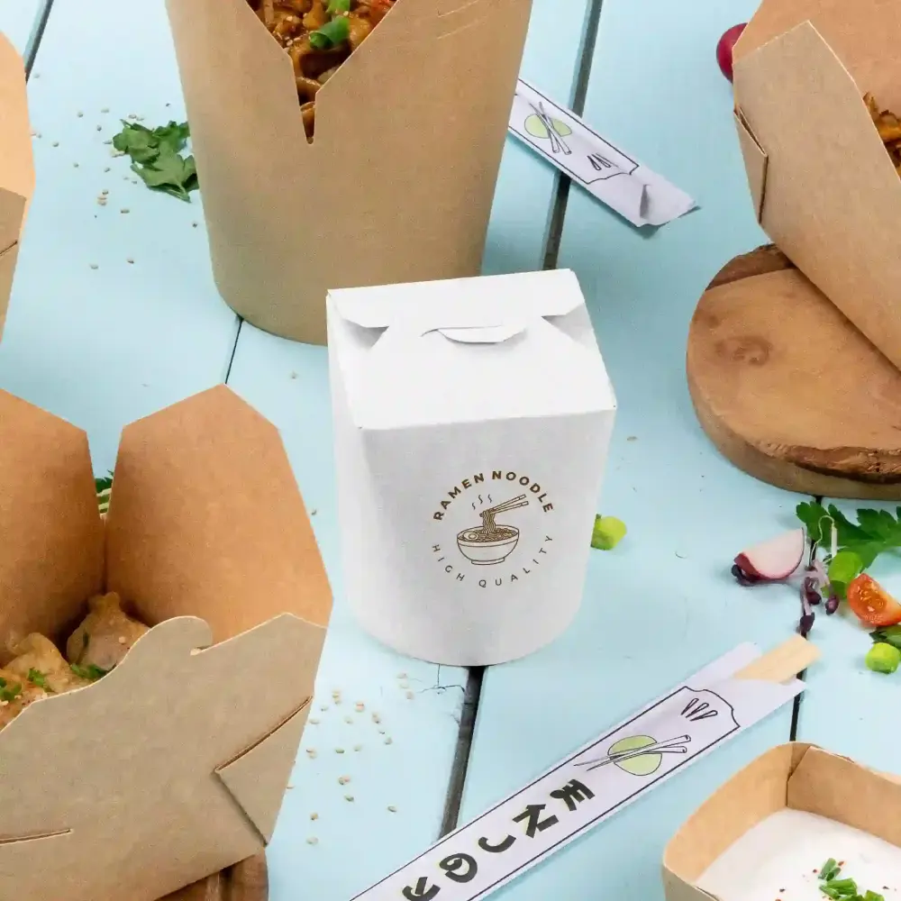 Biała pudełko na wynos z napisem "Ramen Noodles wysokiej jakości" obok brązowych pudełek na wynos na niebieskiej powierzchni.