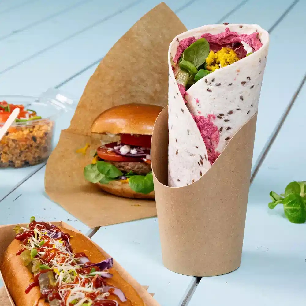 Wrap wypełniony warzywami i pastą, podany w brązowym papierowym pojemniku, w tle hamburger i hot dog.