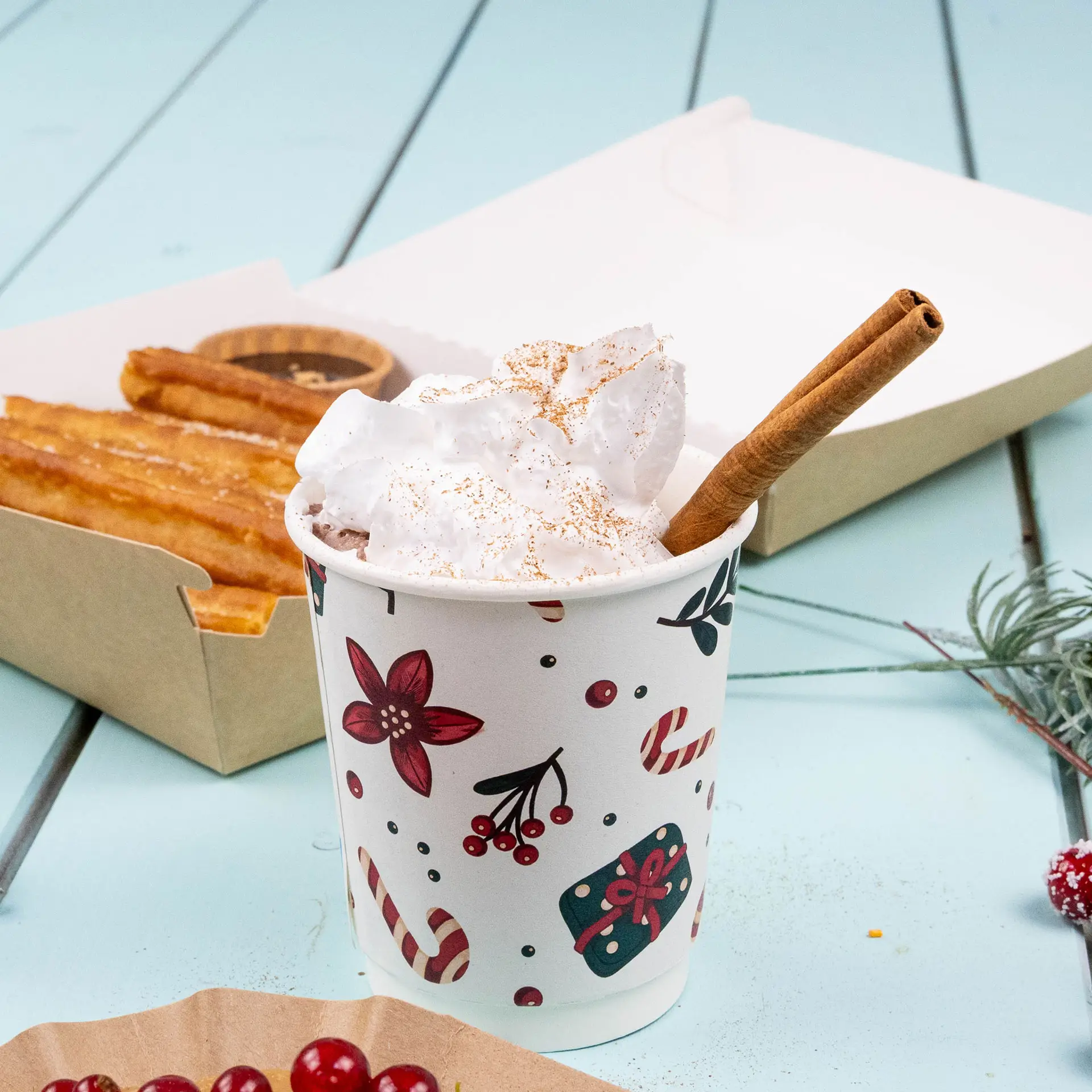 Podwójna szklanka z zimowym wzorem, wypełniona bitą śmietaną i laską cynamonu, obok churros.