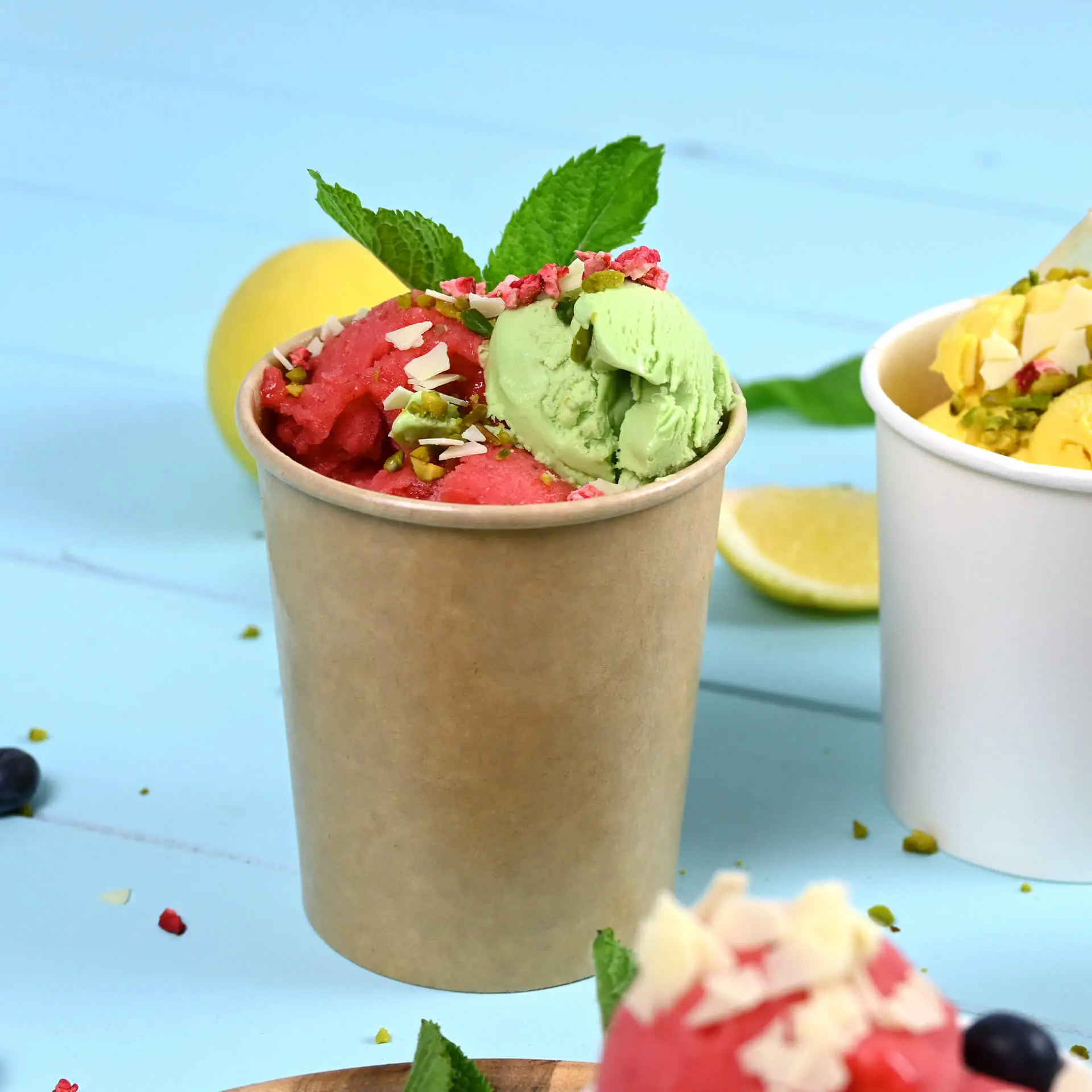 Duży brązowy papierowy kubek na lody wypełniony różowym i zielonym lodem, posypany orzechami i liśćmi mięty, na jasnoniebieskim tle.