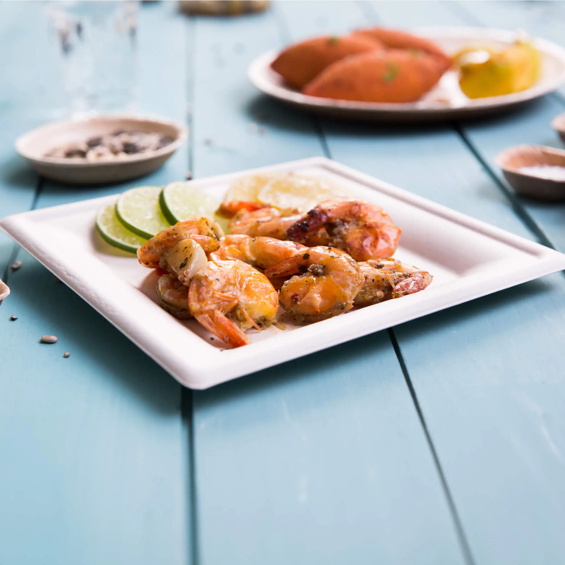 Grillowane krewetki z plasterkami limonki podane na kwadratowym talerzu z trzciny cukrowej; dodatkowe jedzenie widoczne w tle.