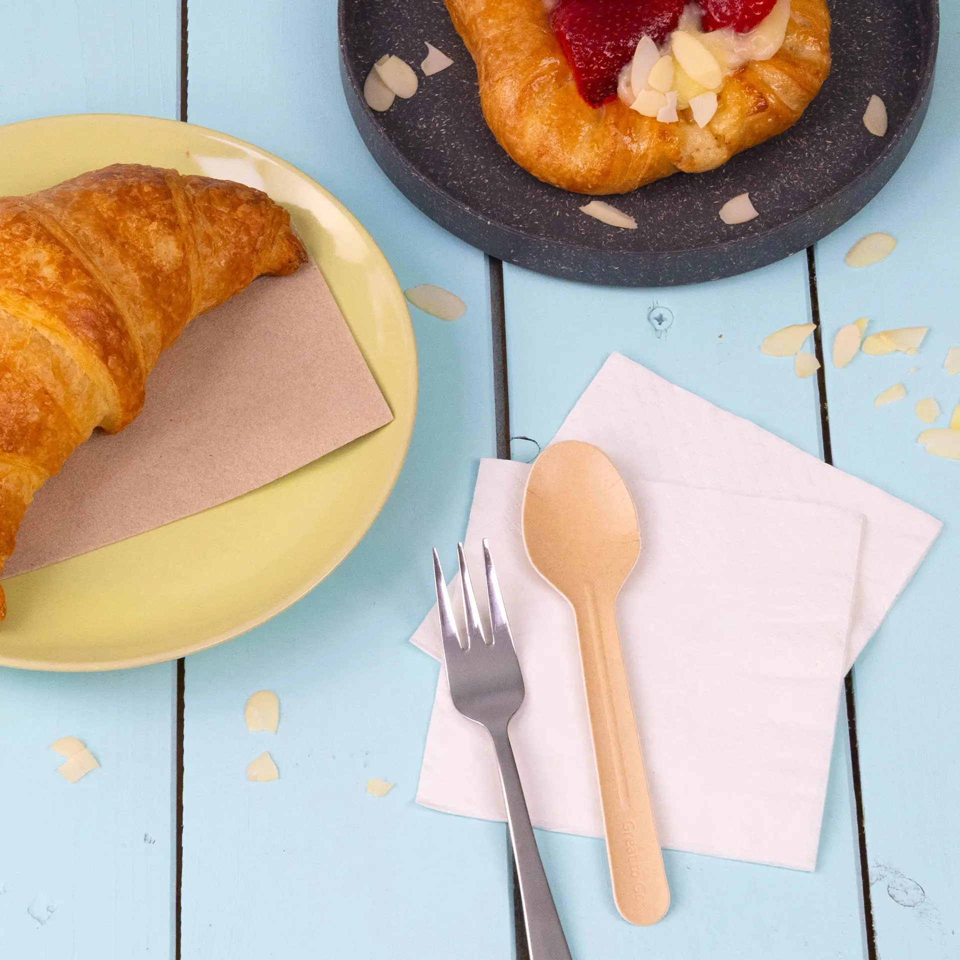 Obraz croissanta na żółtym talerzu, ciastka na ciemnym talerzu, drewniana łyżka, srebrny widelec i białe serwetki.
