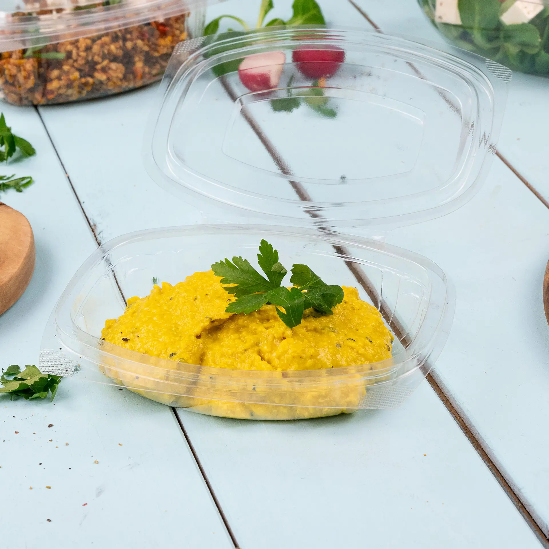 Miska gastronomiczna z żółtym dipem, ozdobiona natką pietruszki; przezroczysta pokrywka częściowo uniesiona, otoczona składnikami sałatki.