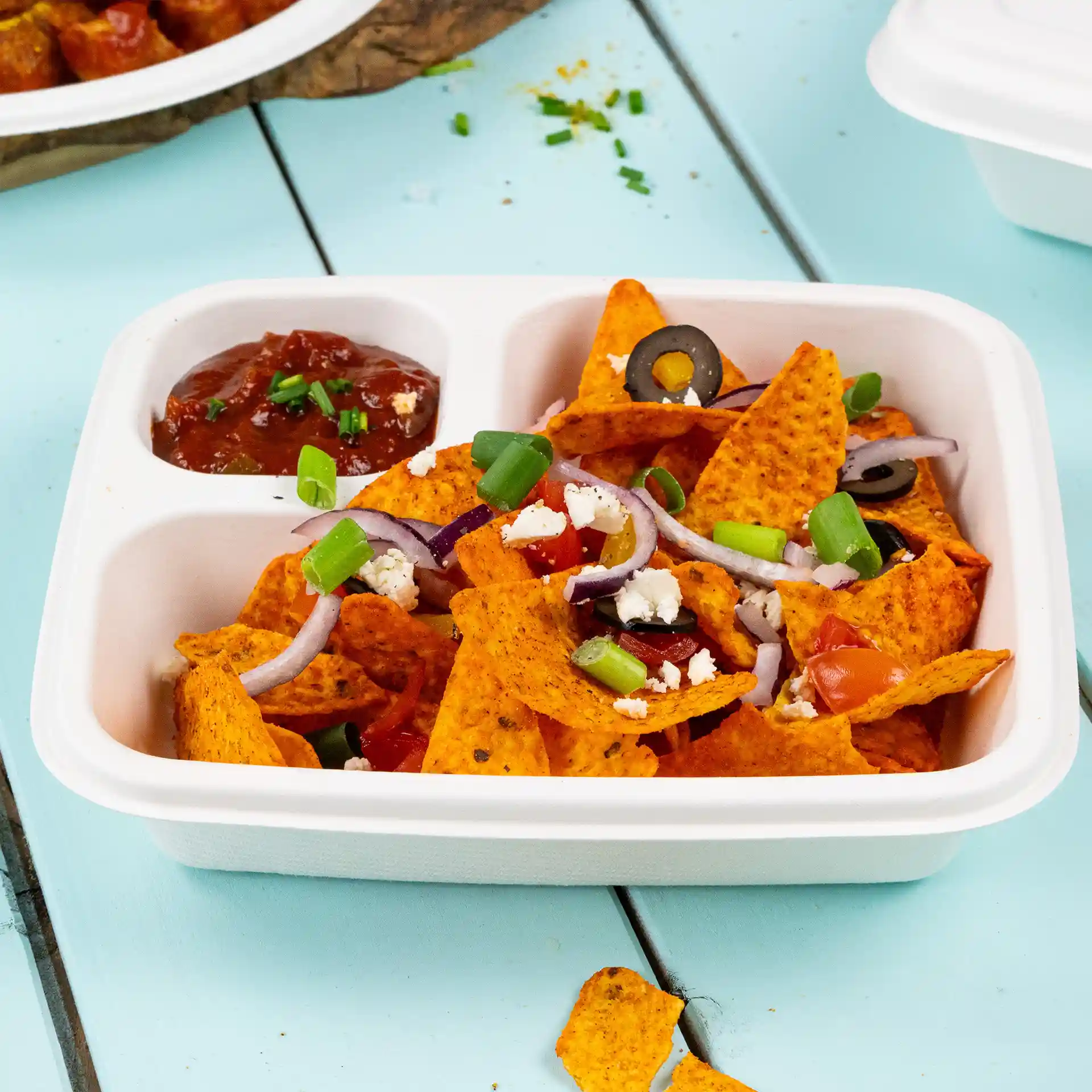 Prostokątny biały talerz z trzciny cukrowej wypełniony nachos, z pomidorami, oliwkami, cebulą i szczypiorkiem, z salsą obok.