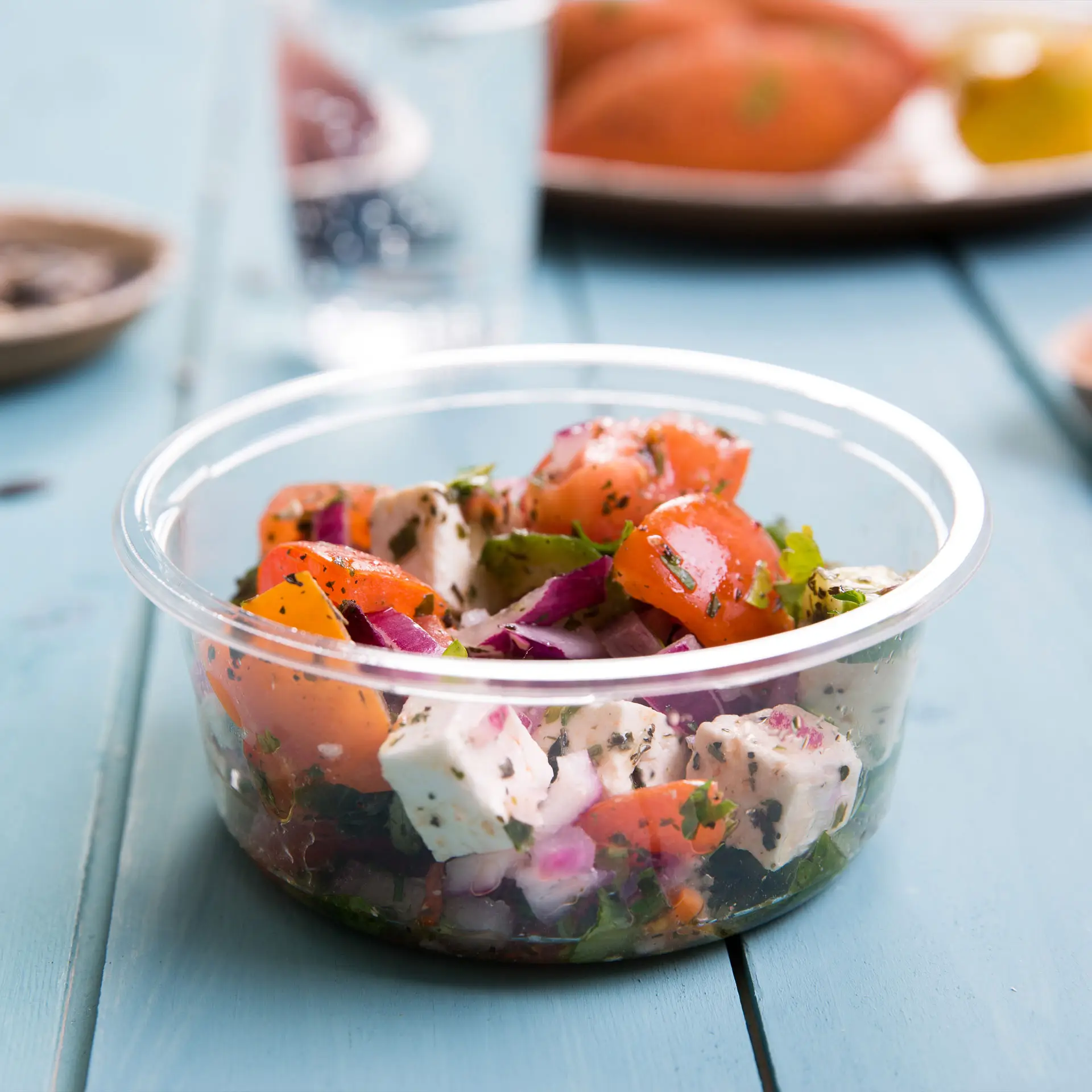 Przezroczysty pojemnik deli z PLA wypełniony świeżymi składnikami sałatkowymi, w tym pomidorami, czerwonymi cebulami i ziołami.