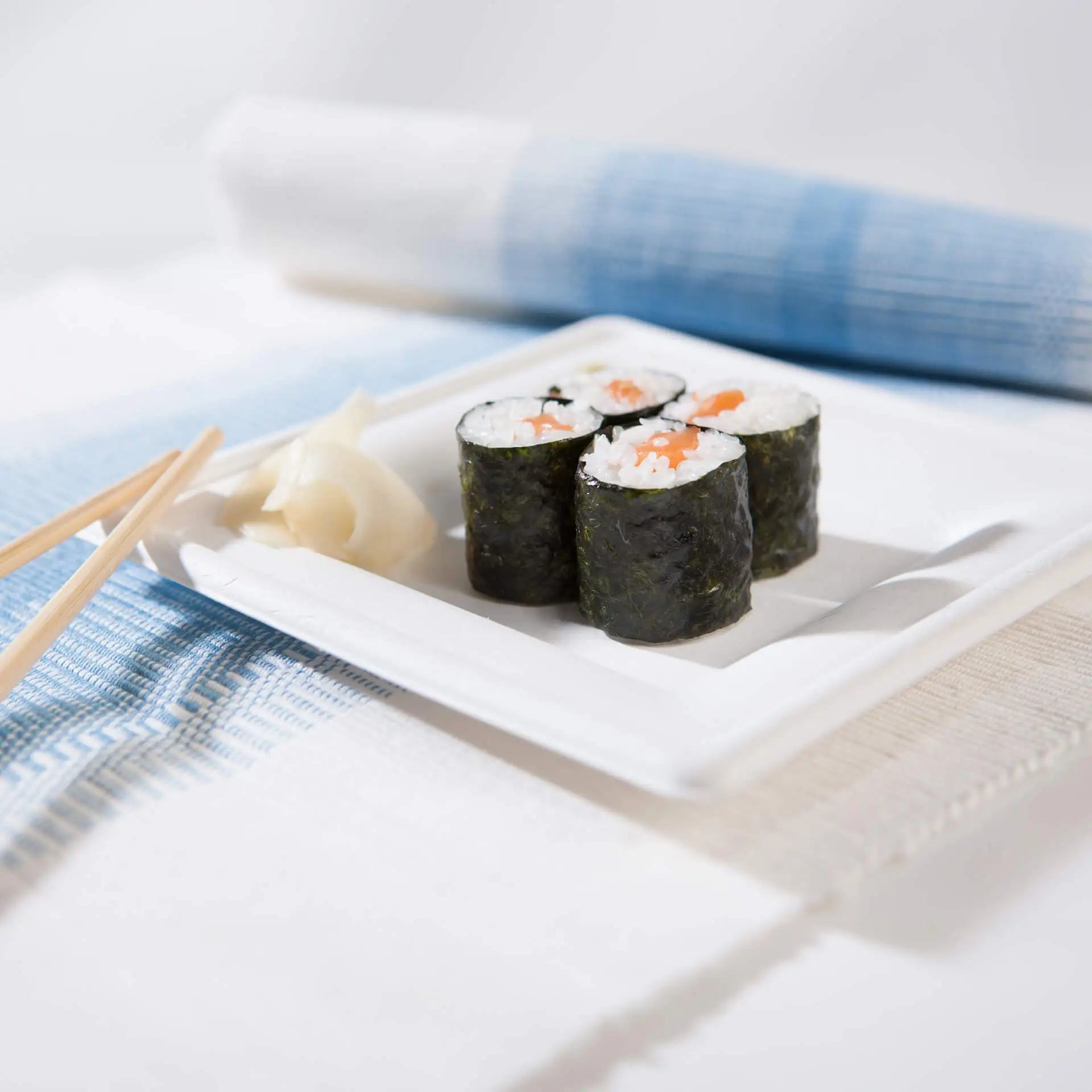 Kwadratowy talerz z trzciny cukrowej z rolkami sushi i marynowanym imbirem, na niebiesko-białym nakryciu stołu.