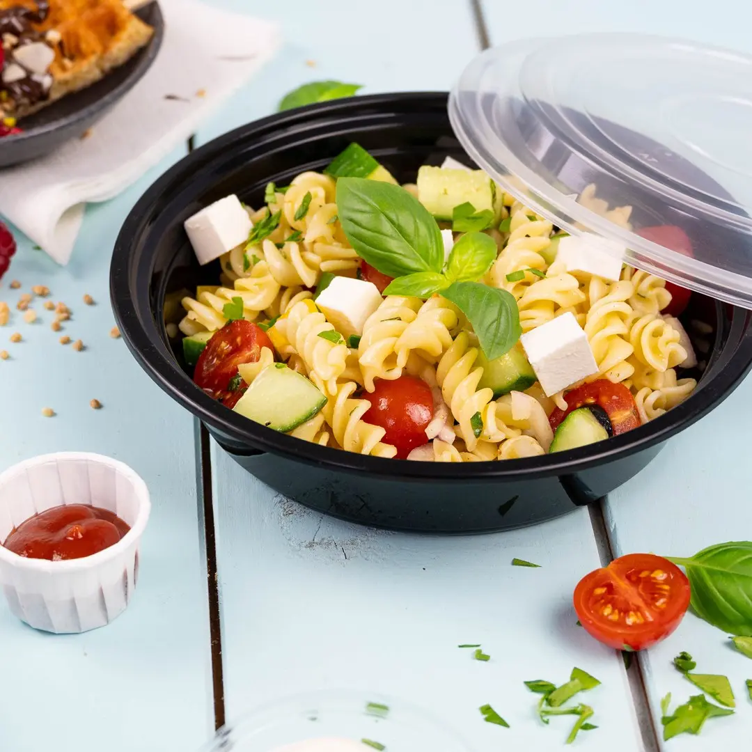 Czarna miska z sałatką makaronową z rotini, pomidorkami koktajlowymi, ogórkiem, serem feta i bazylią, z ketchupem.