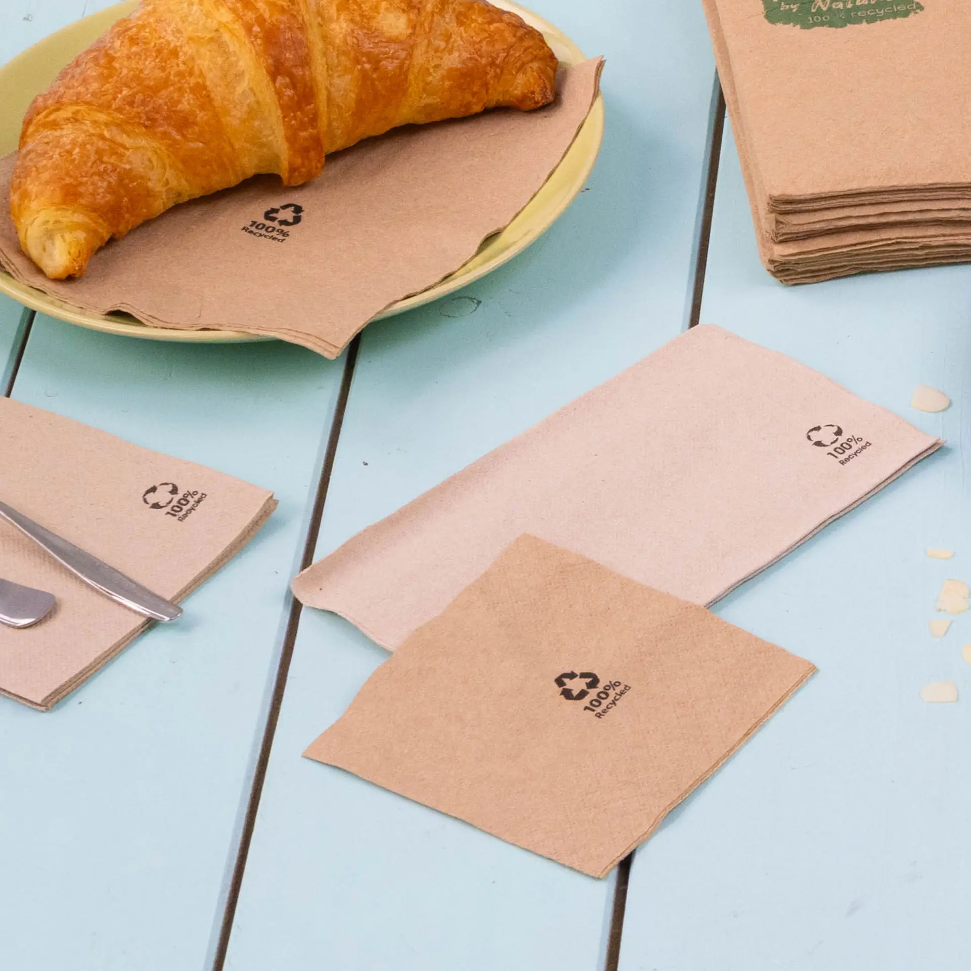 Obraz brązowych, biodegradowalnych serwetek koktajlowych na niebieskim stole, z rogalikiem na talerzu, prezentujących symbole recyklingu.