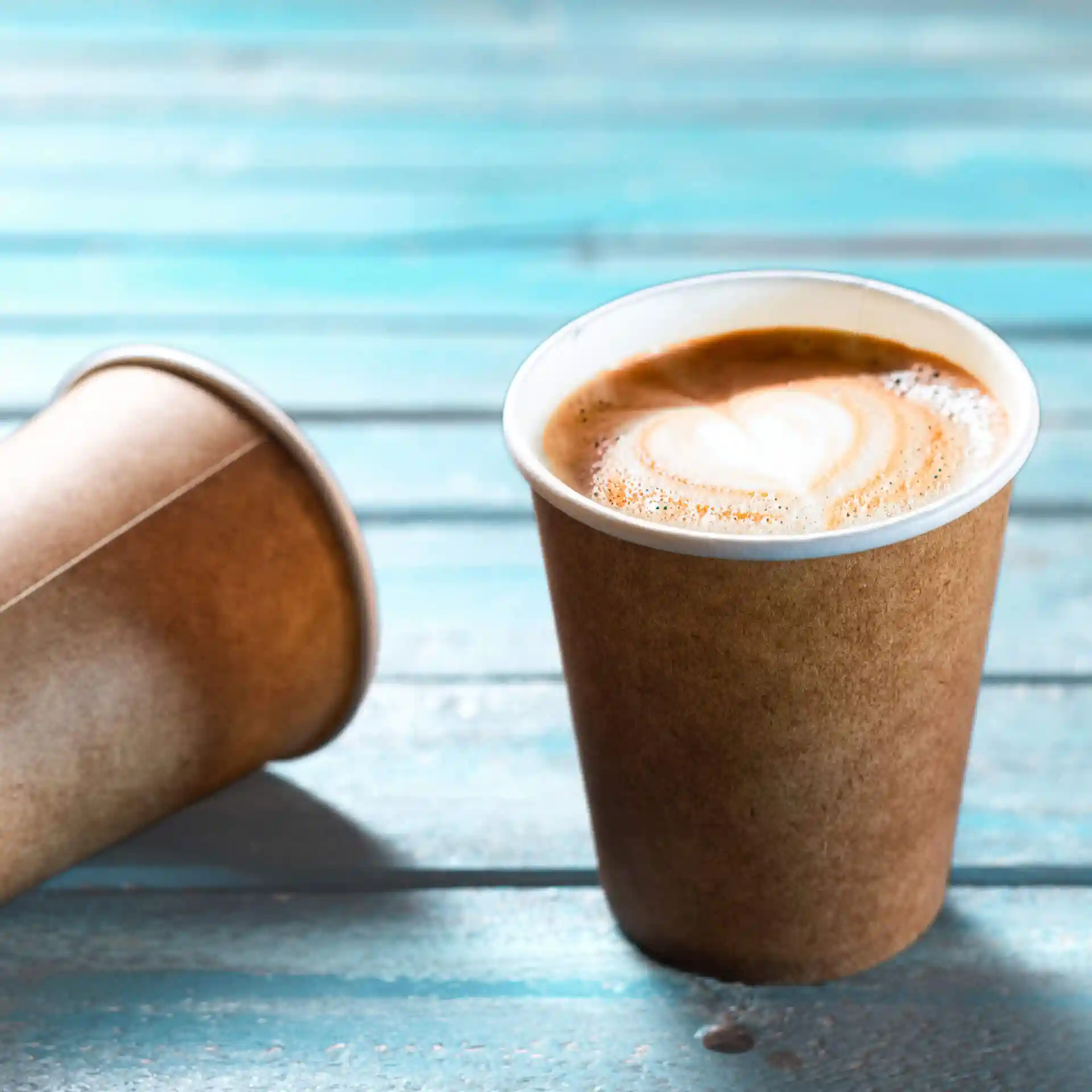 Brąski kubek papierowy z białym wnętrzem, wypełniony kawą i latte art w kształcie serca, na niebieskiej drewnianej powierzchni.