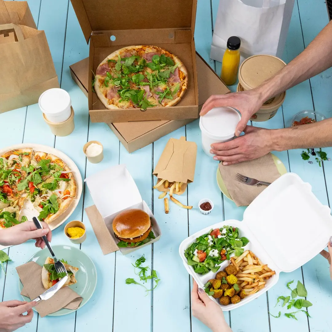 Stół nakryty różnymi daniami na wynos, w tym pizzami, burgerem, frytkami, sałatką i napojami w pojemnikach.