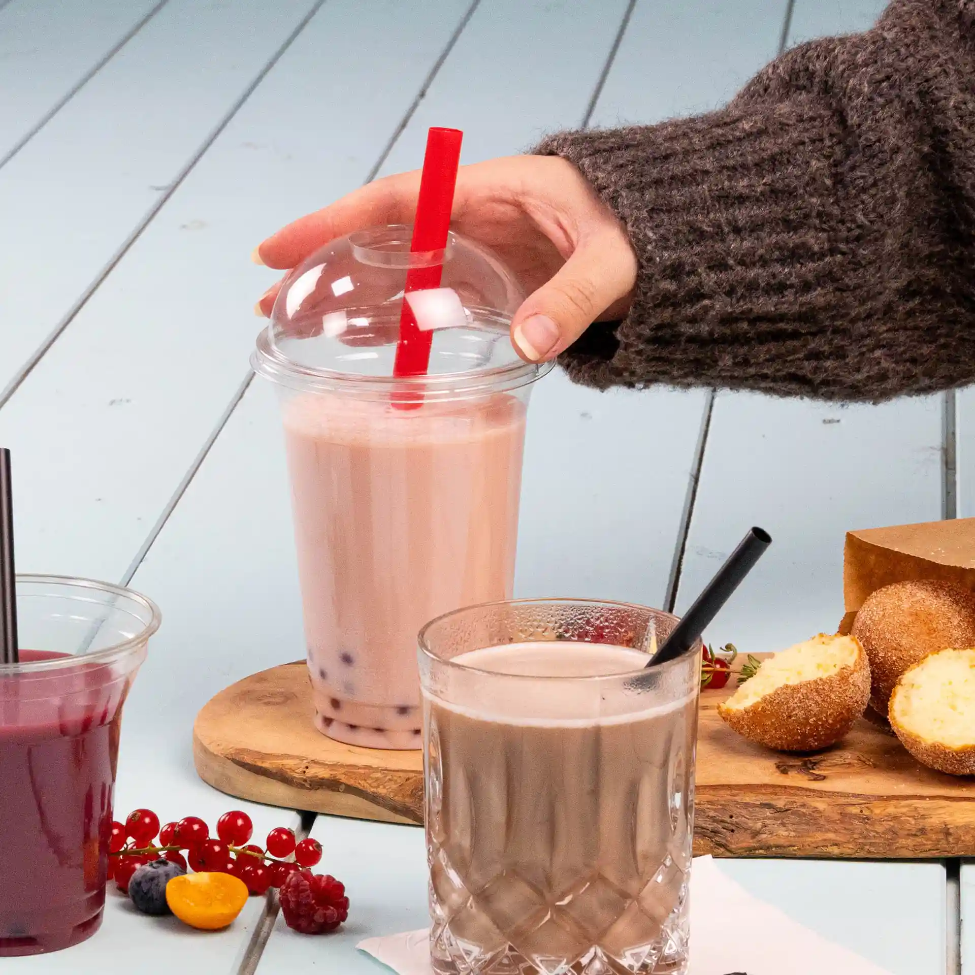 Obraz ręki trzymającej kubek bubble tea z czerwoną słomką, obok szklanki napoju czekoladowego i przekąsek na drewnianej desce.