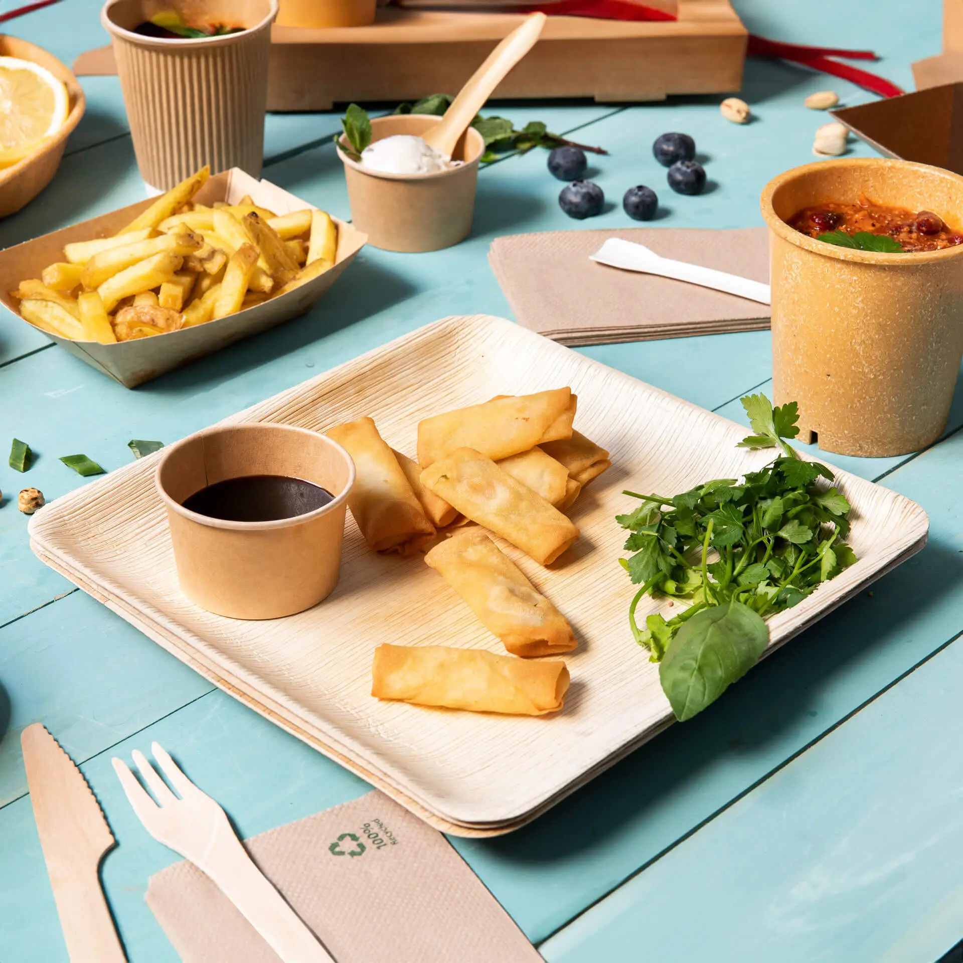 Jedna kwadratowa talerz z liścia palmowego z sajgonkami, miseczką sosu i świeżymi ziołami; podane na niebieskim stole z dodatkami.