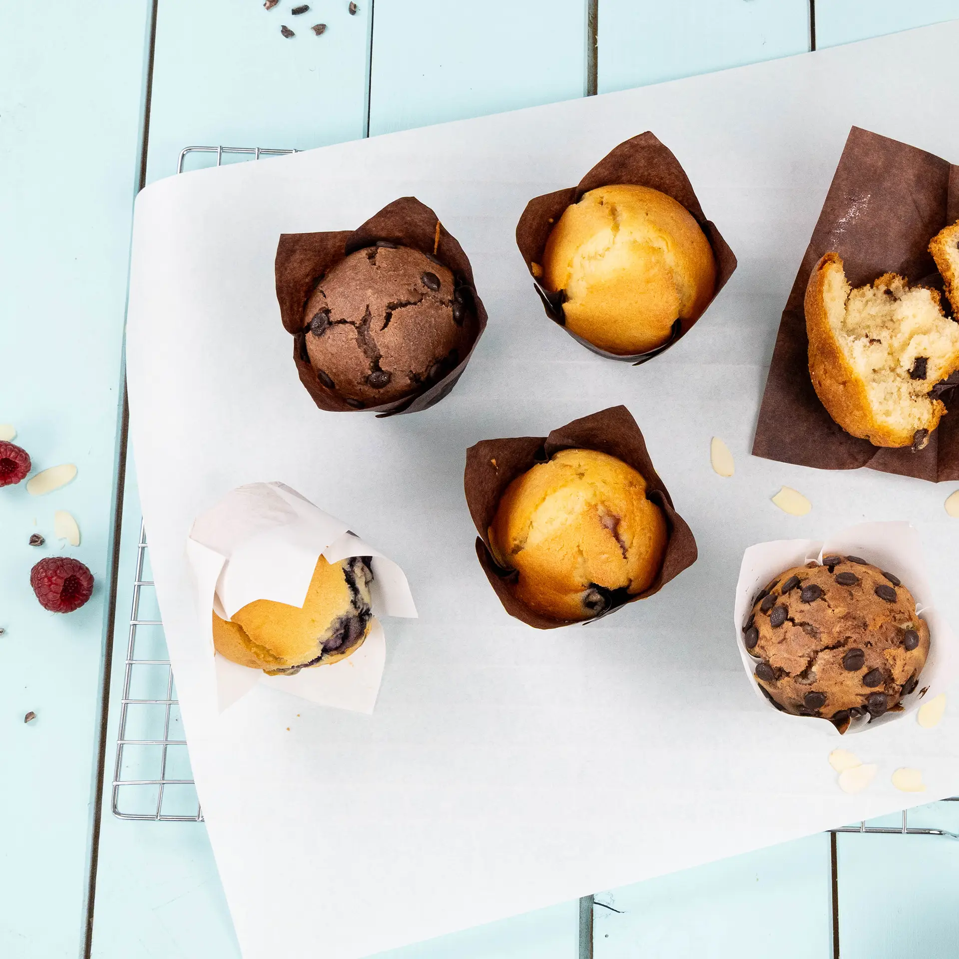 Różne muffiny w papierowych kubkach na białej blaszce do pieczenia, obok kilka malin i płatków migdałowych.