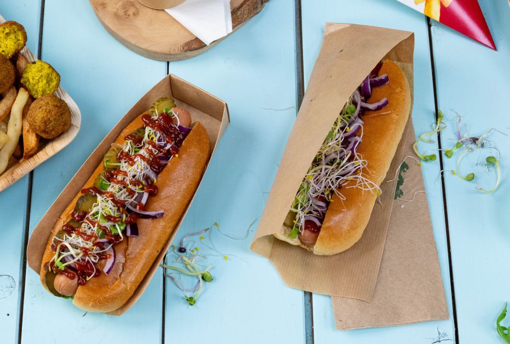 Dwa hot dogi z różnymi dodatkami, w tym ogórkami, cebulą i kiełkami, podane w papierowych pojemnikach na niebieskim stole.