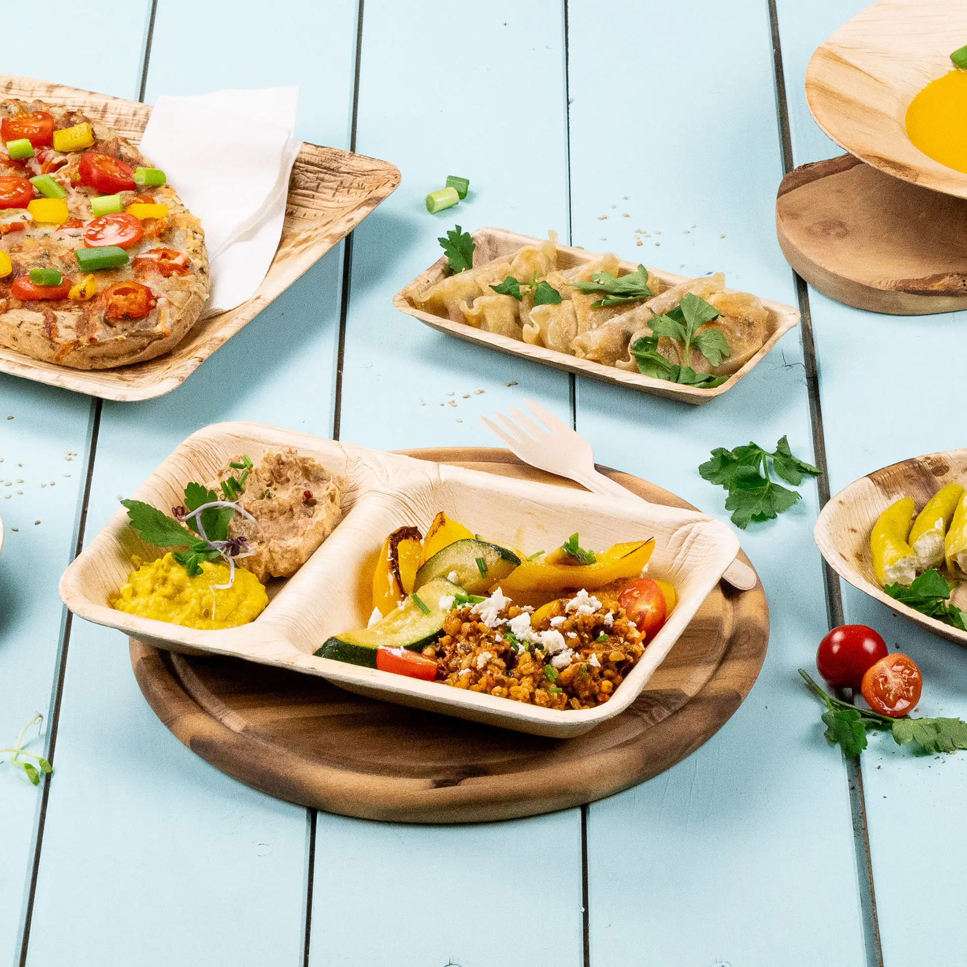 Talerz menu z trzema przegródkami z liści palmowych z hummusem, duszonymi warzywami i pierożkami na jasnoniebieskim tle.