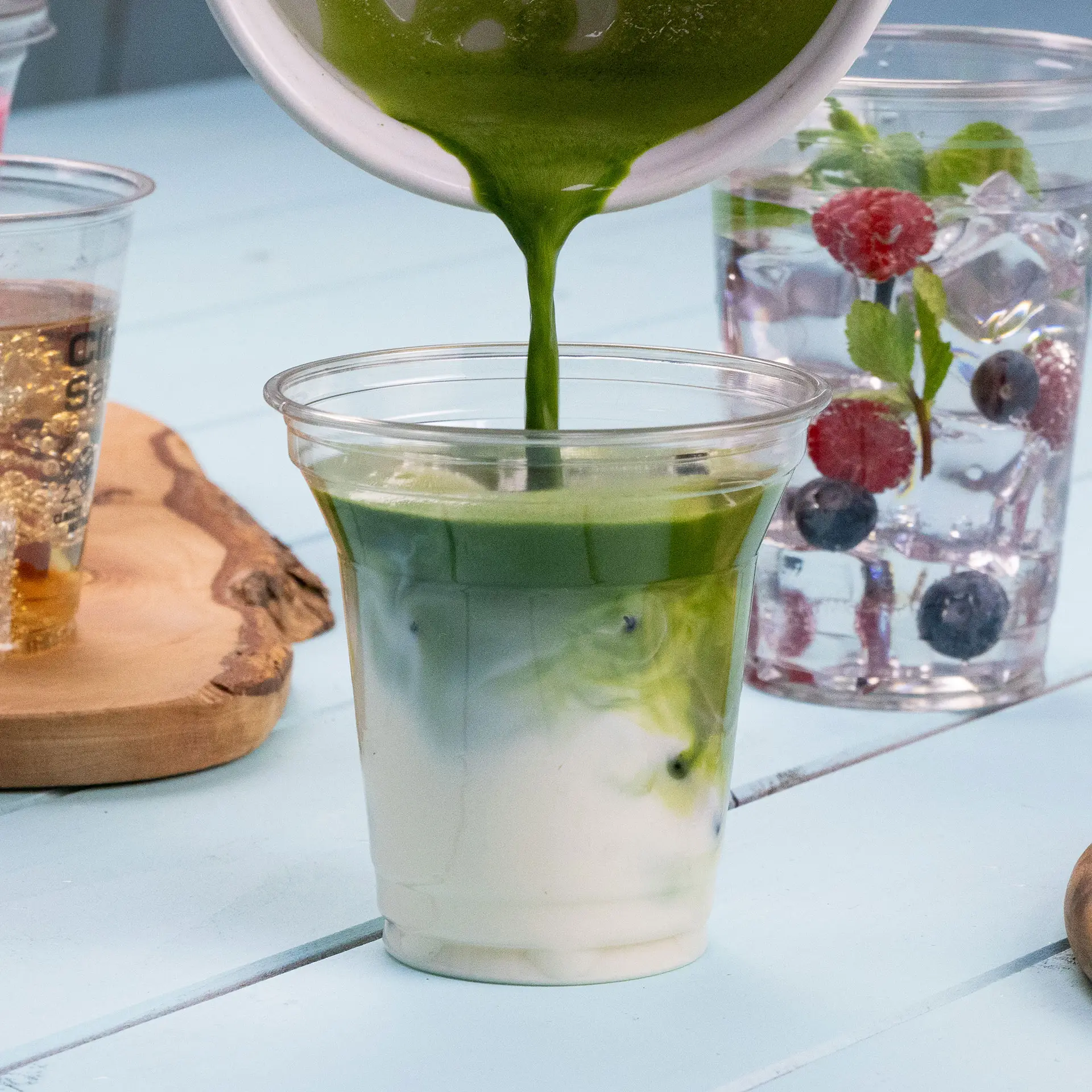 Przezroczysty kubek rPET jest napełniany zielonym napojem, widoczne warstwy koloru; w tle widać dodatkowe napoje.