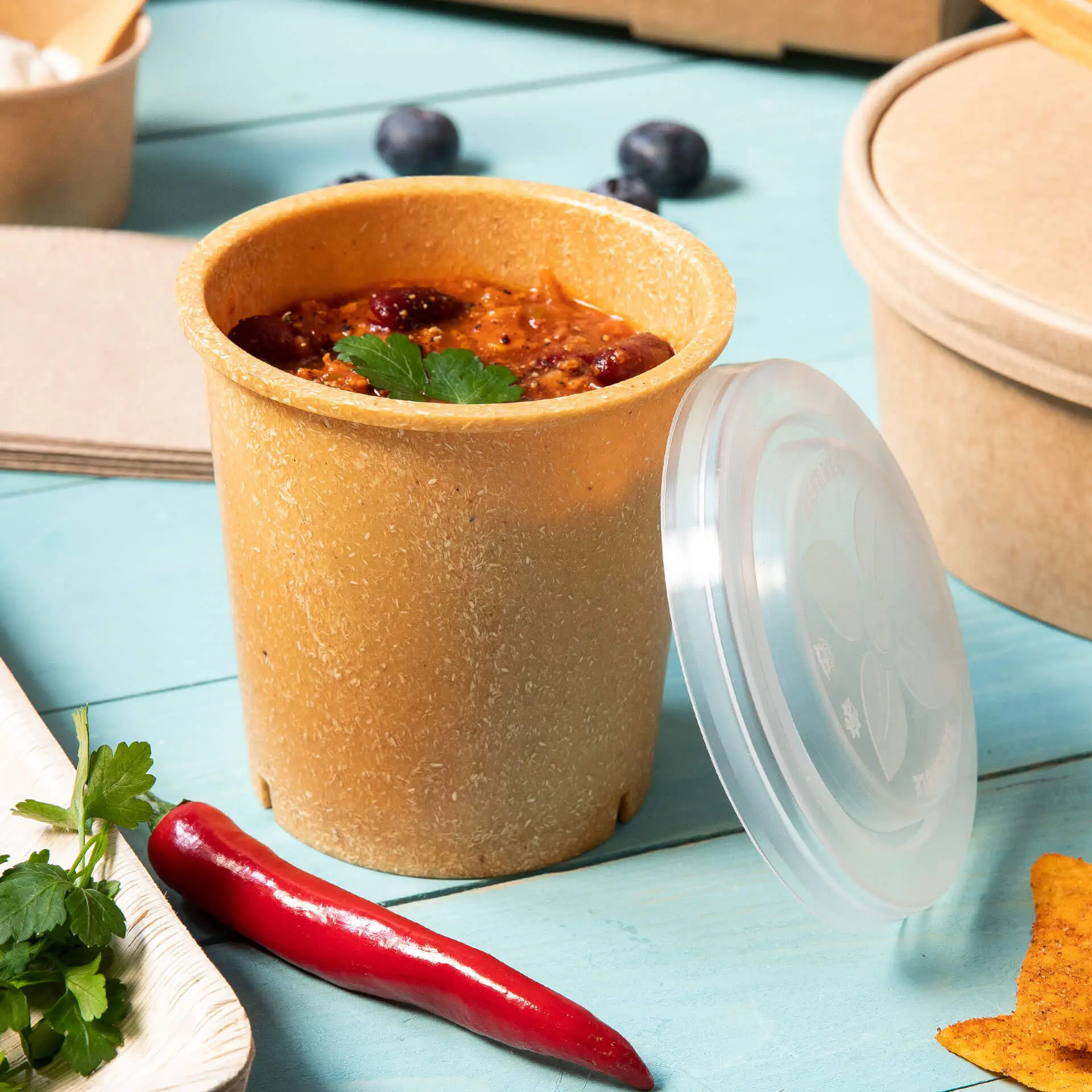 Jedna karamelowa wielorazowa miseczka na zupę z pokrywką, wypełniona zupą i przyozdobiona kolendrą; obok czerwona papryczka chili.