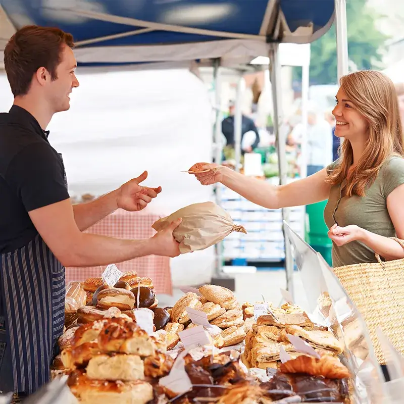Sprzedawca wręcza brązową torbę papierową klientowi na stoisku z różnymi wypiekami.