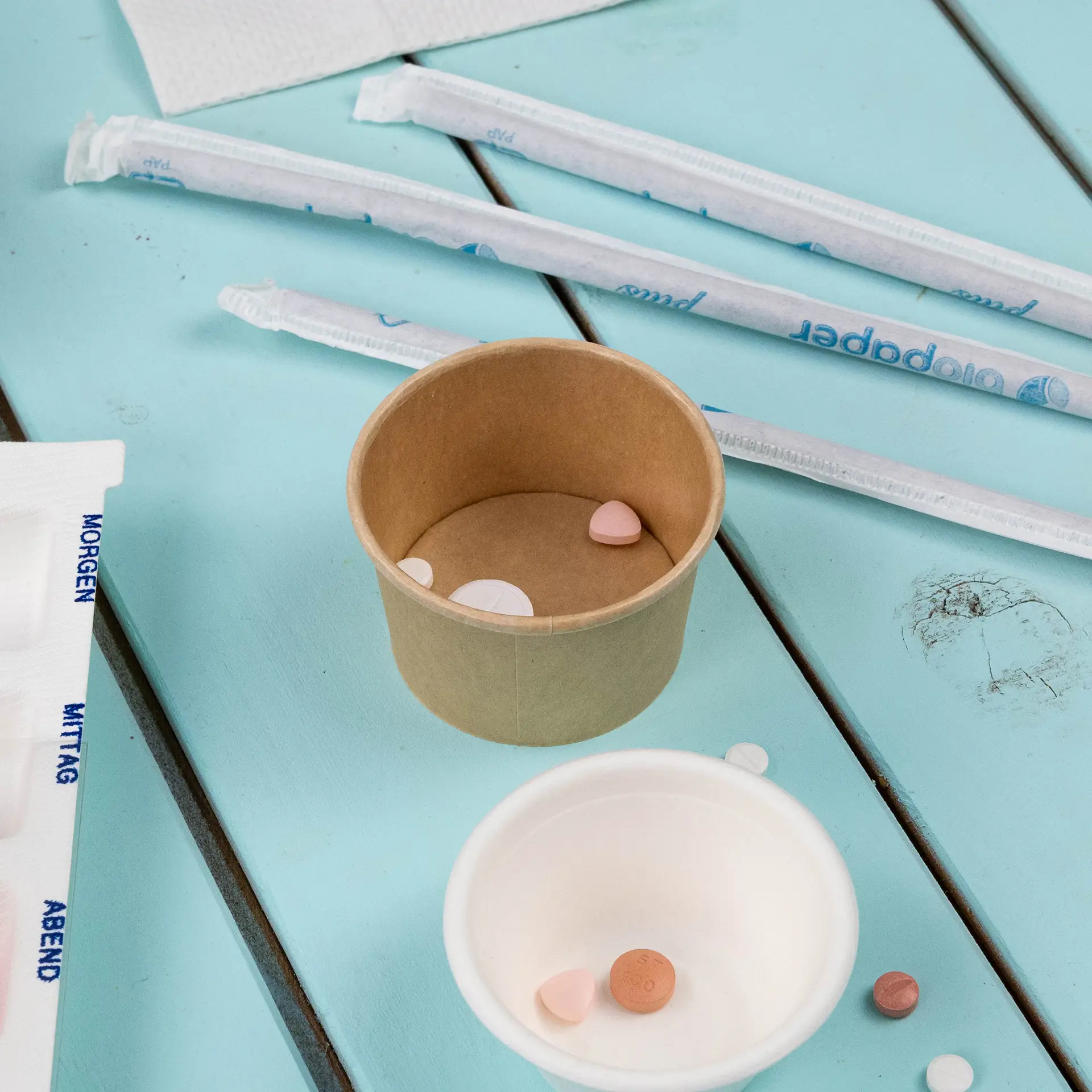 Pojemnik Deli (brązowy, 40 ml) z małymi kolorowymi pigułkami w środku; dodatkowe pojemniki i słomki są obok na niebieskiej powierzchni.