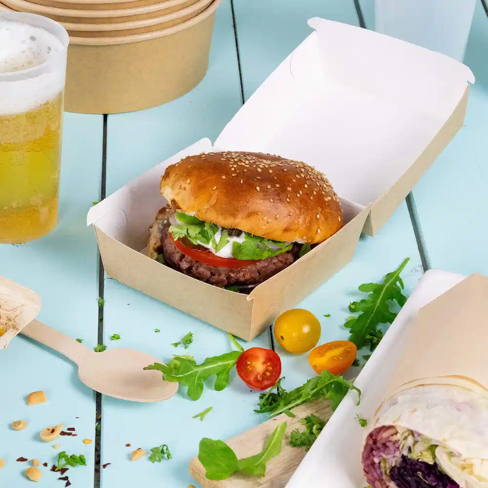 Burger z bułką sezamową, sałatą, pomidorem i sosem w brązowym pudełku na wynos, z warzywami i piwem.