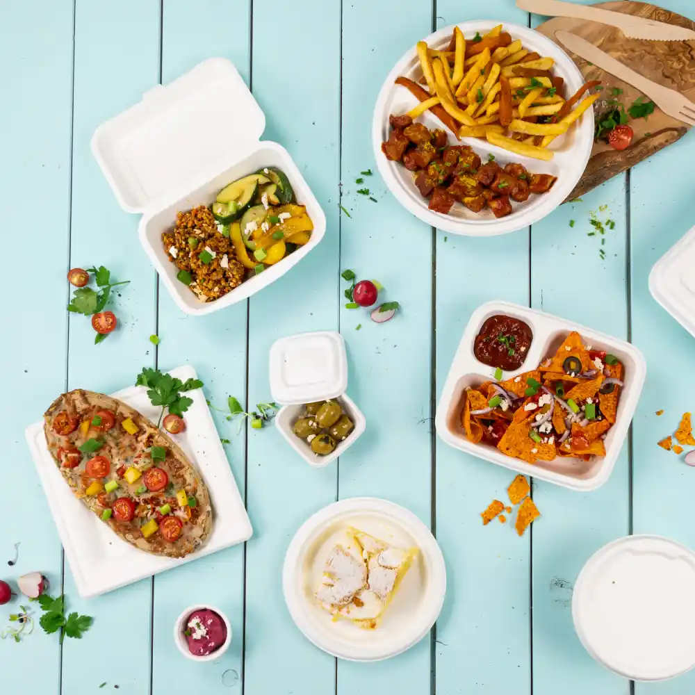 Różnorodne jednorazowe pojemniki na żywność z różnymi potrawami, w tym frytkami, nachos, sałatką i deserem na niebieskim drewnianym stole.