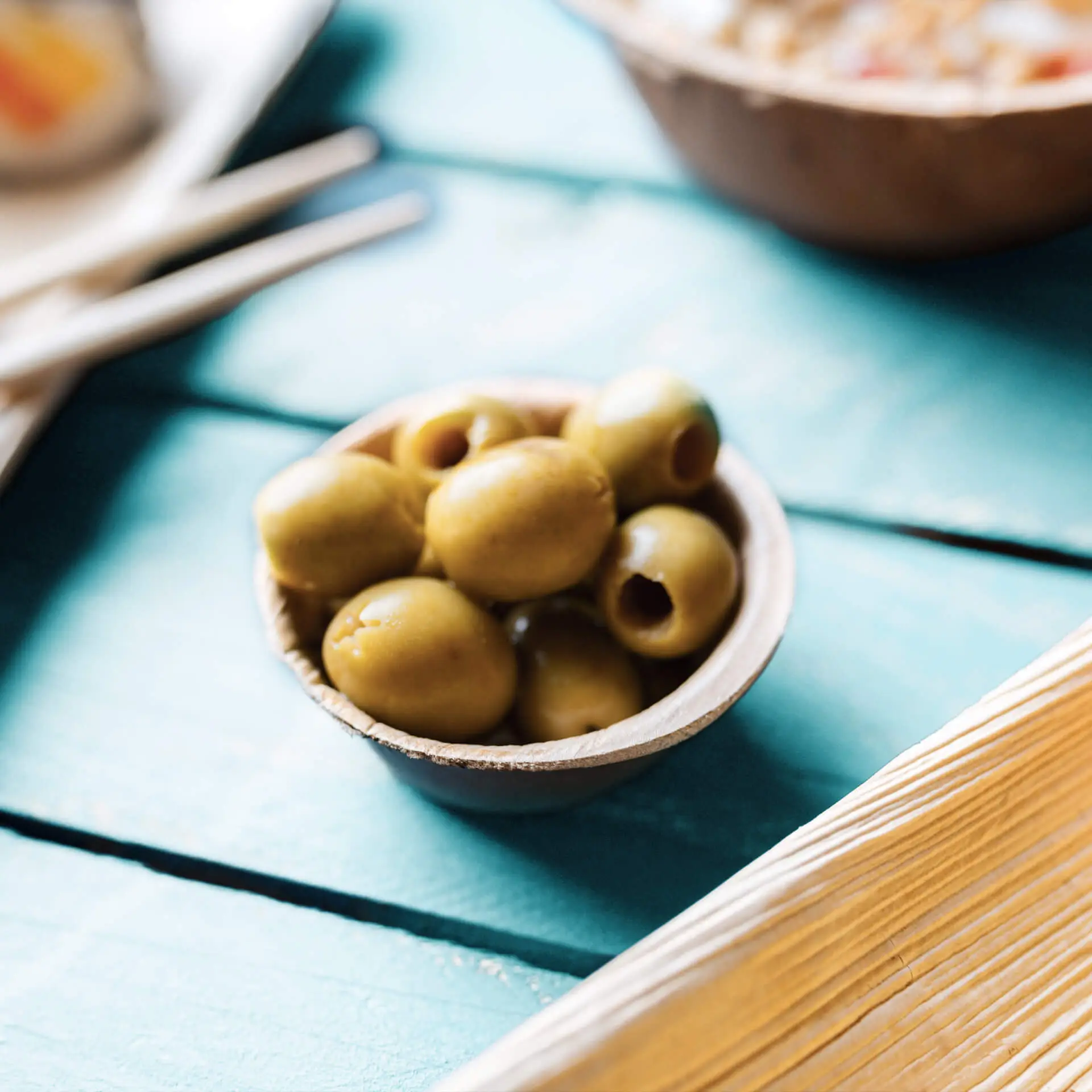 Mała okrągła miska z liści palmowych wypełniona zielonymi oliwkami, ustawiona na turkusowej drewnianej powierzchni.