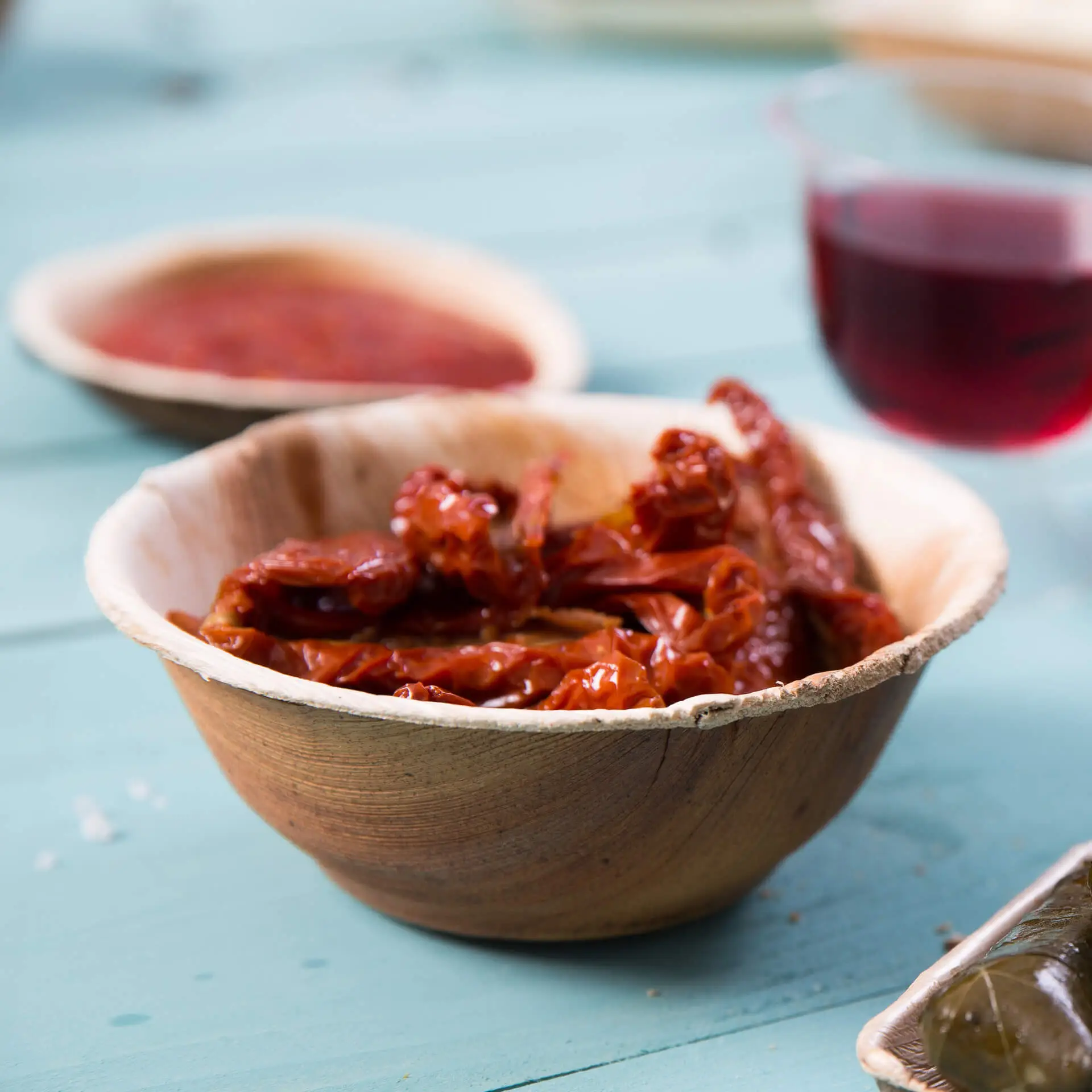 Miska z liści palmowych wypełniona suszonymi pomidorami; dodatkowa miska i szklanka czerwonego płynu w tle.