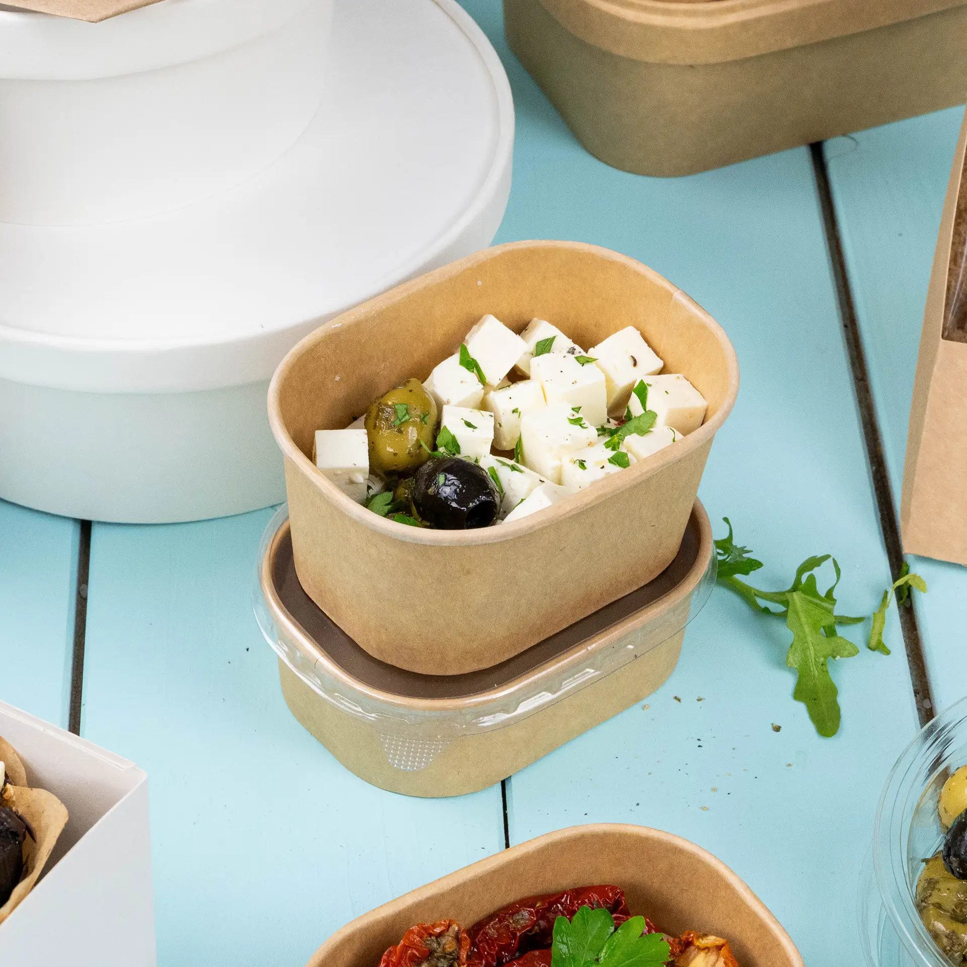 Prostokątny pojemnik kraft na żywność wypełniony kostkami sera i oliwkami, z zieloną dekoracją; widoczne są dodatkowe pojemniki.