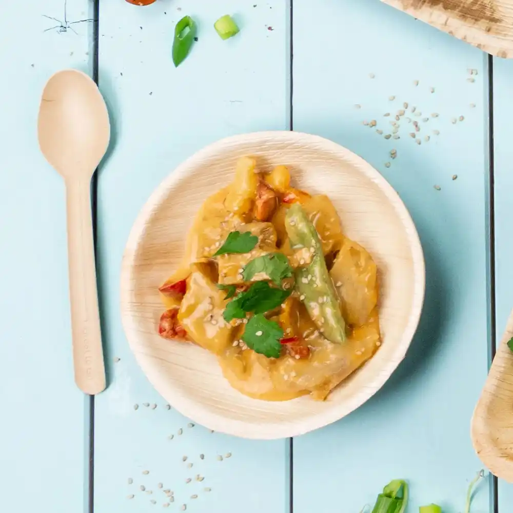 Bamboowa miska z warzywami stir-fry i sosem, posypana nasionami sezamu i kolendrą, obok drewnianej łyżki.