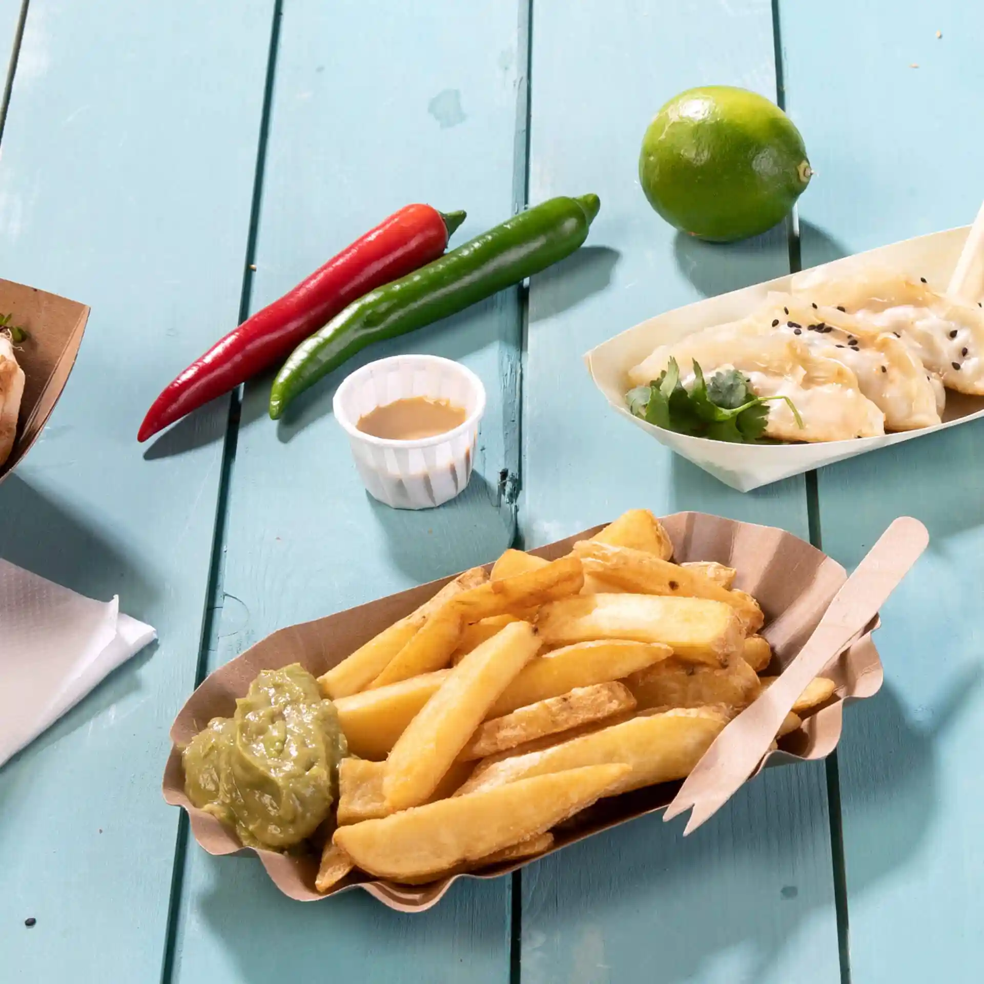 Obraz porcji frytek z zielonym sosem w papierowym pojemniku, otoczonych papryczkami chili, limonką i małym kubkiem sosu.