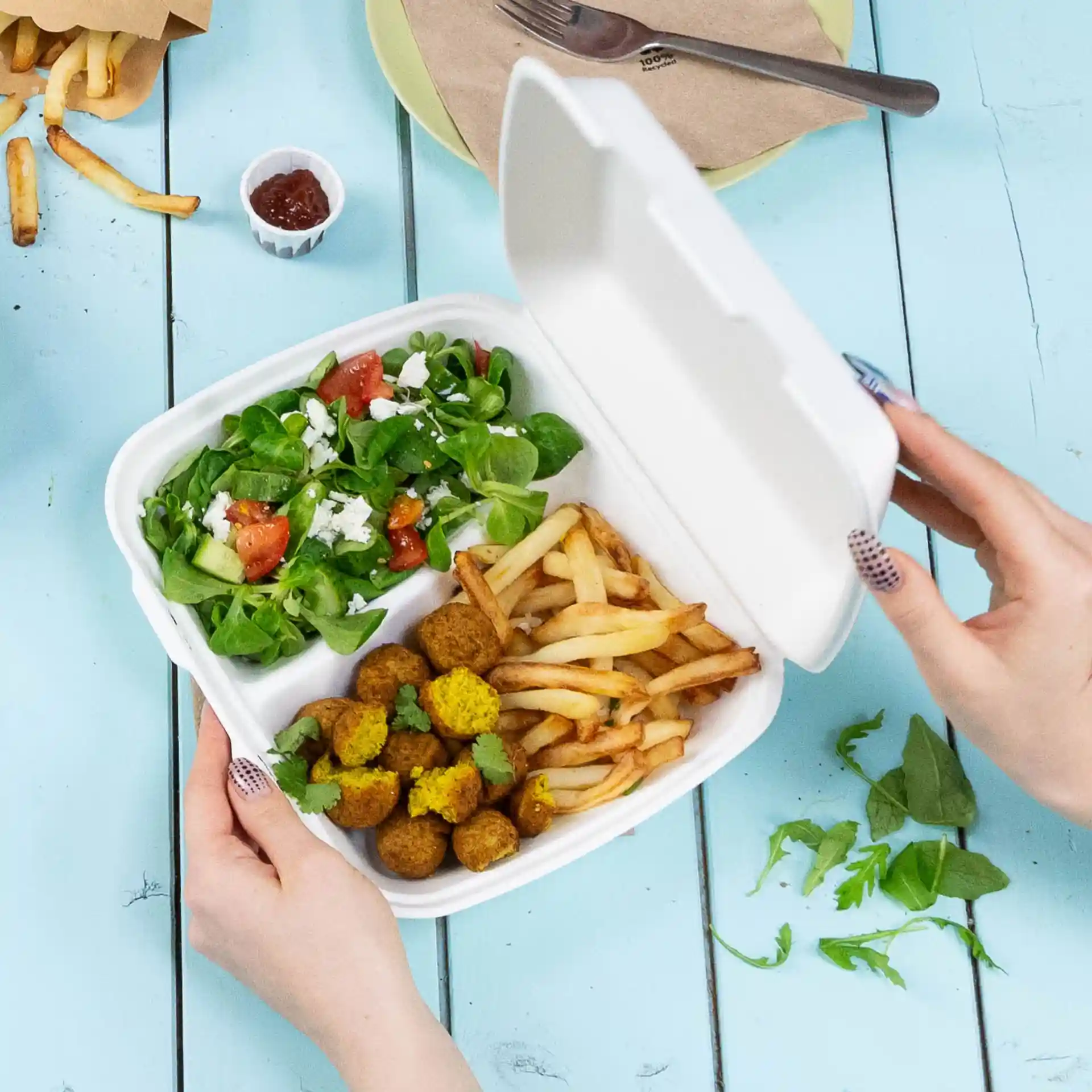 Ręka trzyma biały pojemnik z zawiasową pokrywką z dwoma przegródkami, wypełniony sałatką, frytkami i kulkami falafel.