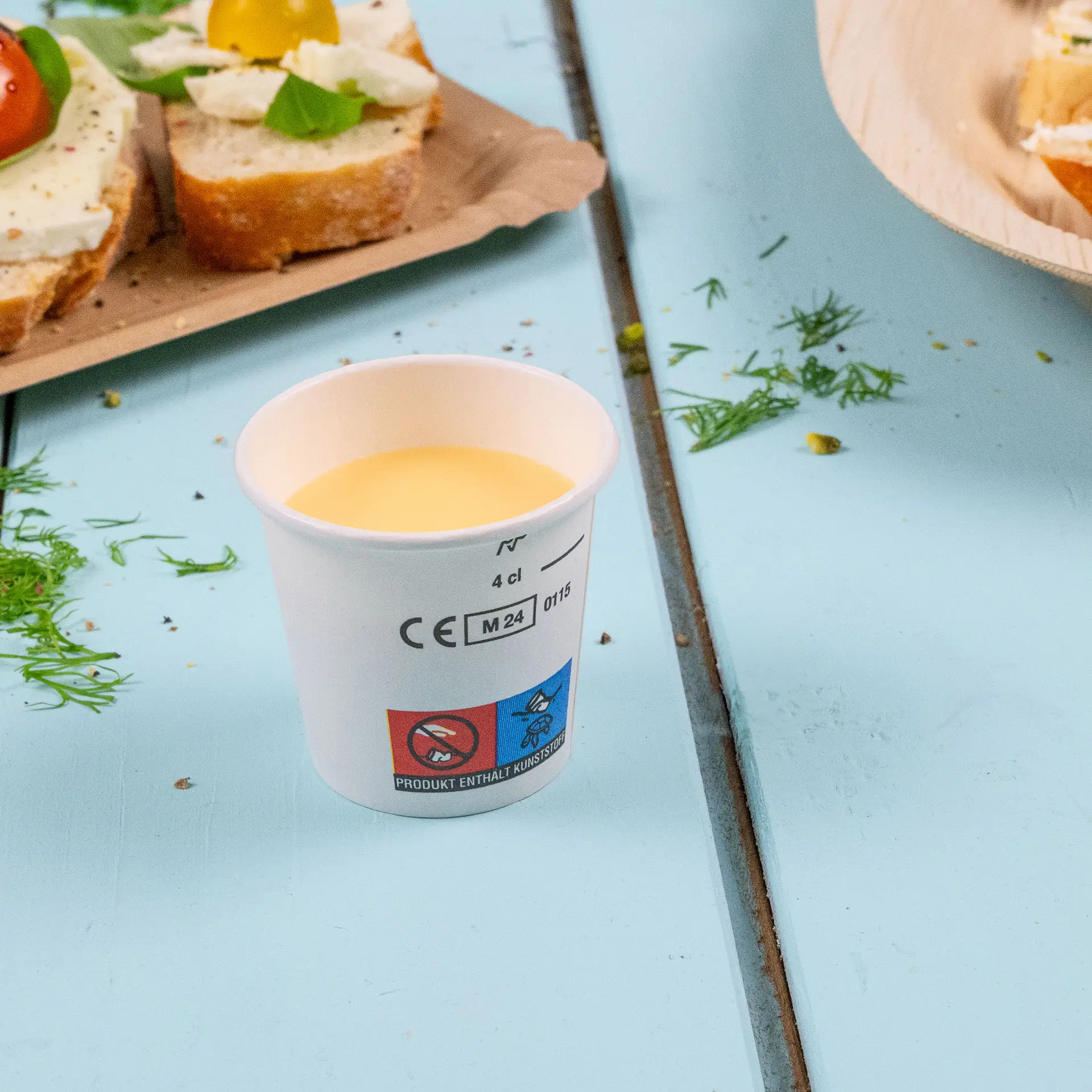 Jedna biała papierowa filiżanka z żółtym płynem; obok znajdują się częściowo widoczne kromki chleba z dodatkami.