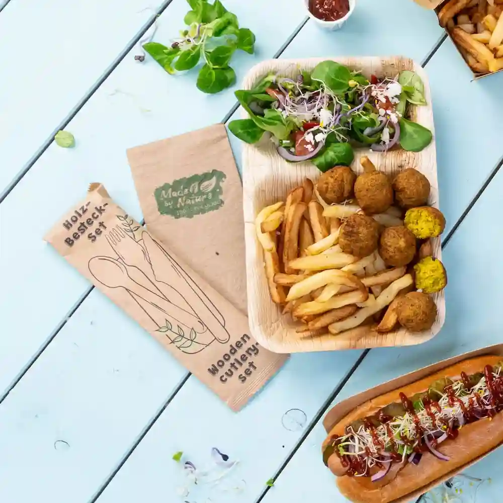 Zestaw drewnianych sztućców obok talerza z jedzeniem, sałatką, frytkami, kulkami falafel i hot dogiem na niebieskim stole.
