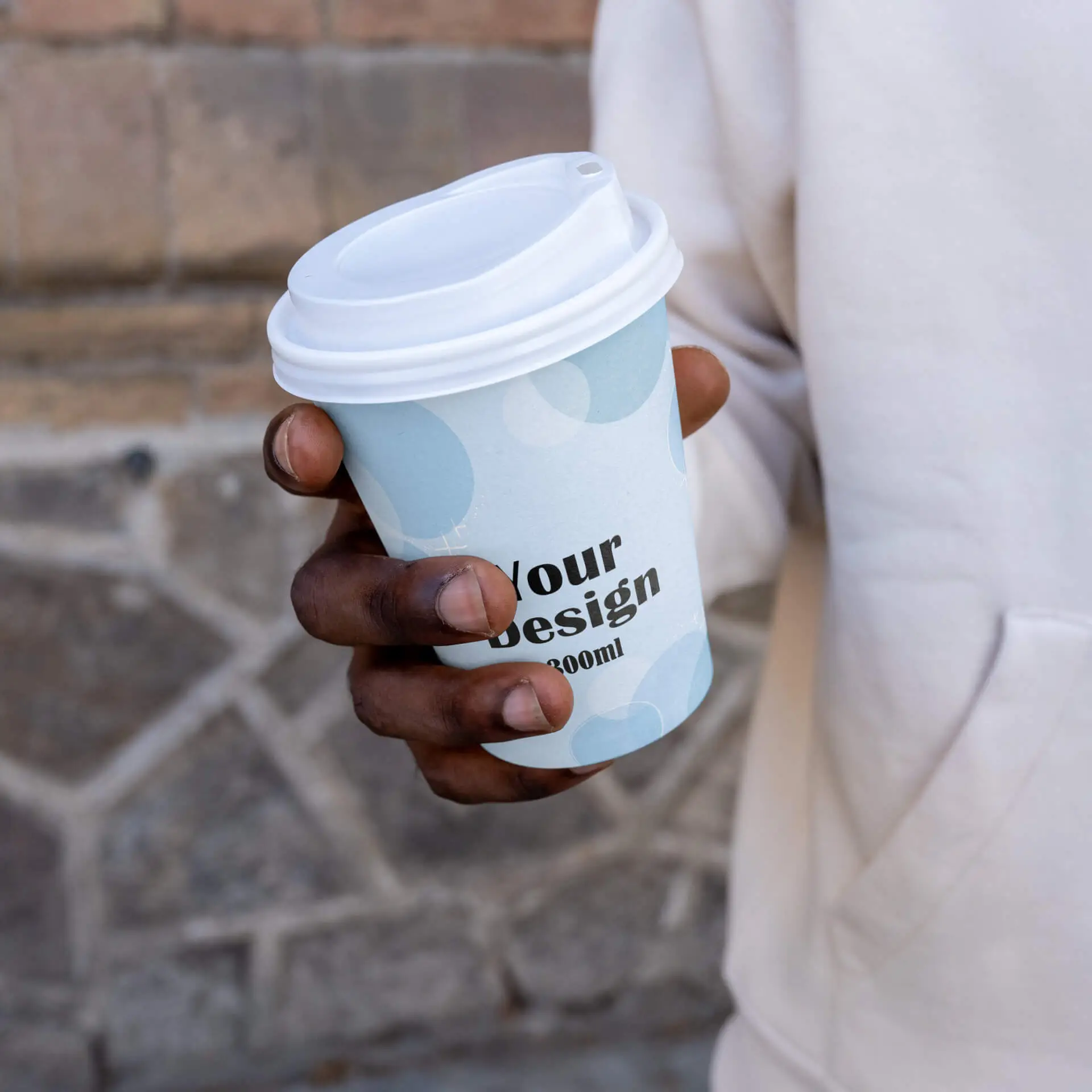 Personalizowany papierowy kubek o pojemności 300 ml w niebieskim wzorze i czarnym tekście; trzymany w dłoni, na tle kamienia.