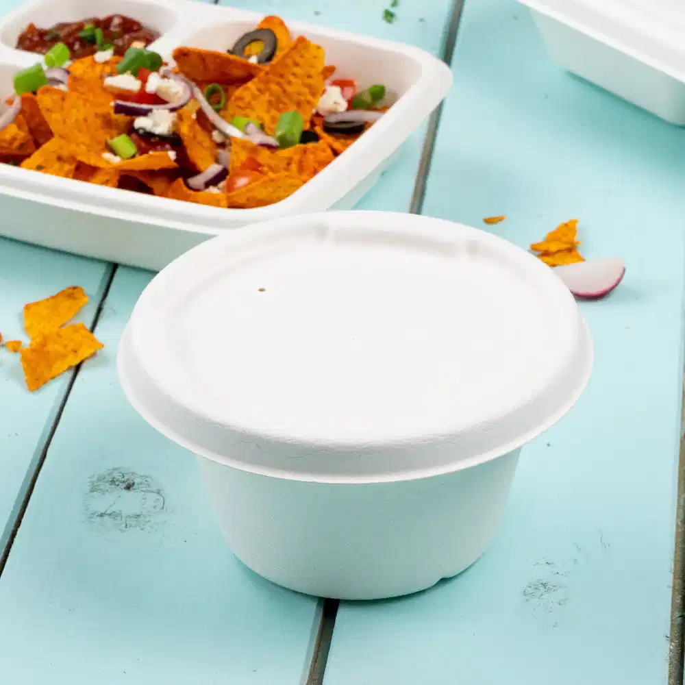 Biały papierowy kubek na zupę na niebieskim stole z tłem nachos i zamkniętym pojemnikiem na jedzenie.