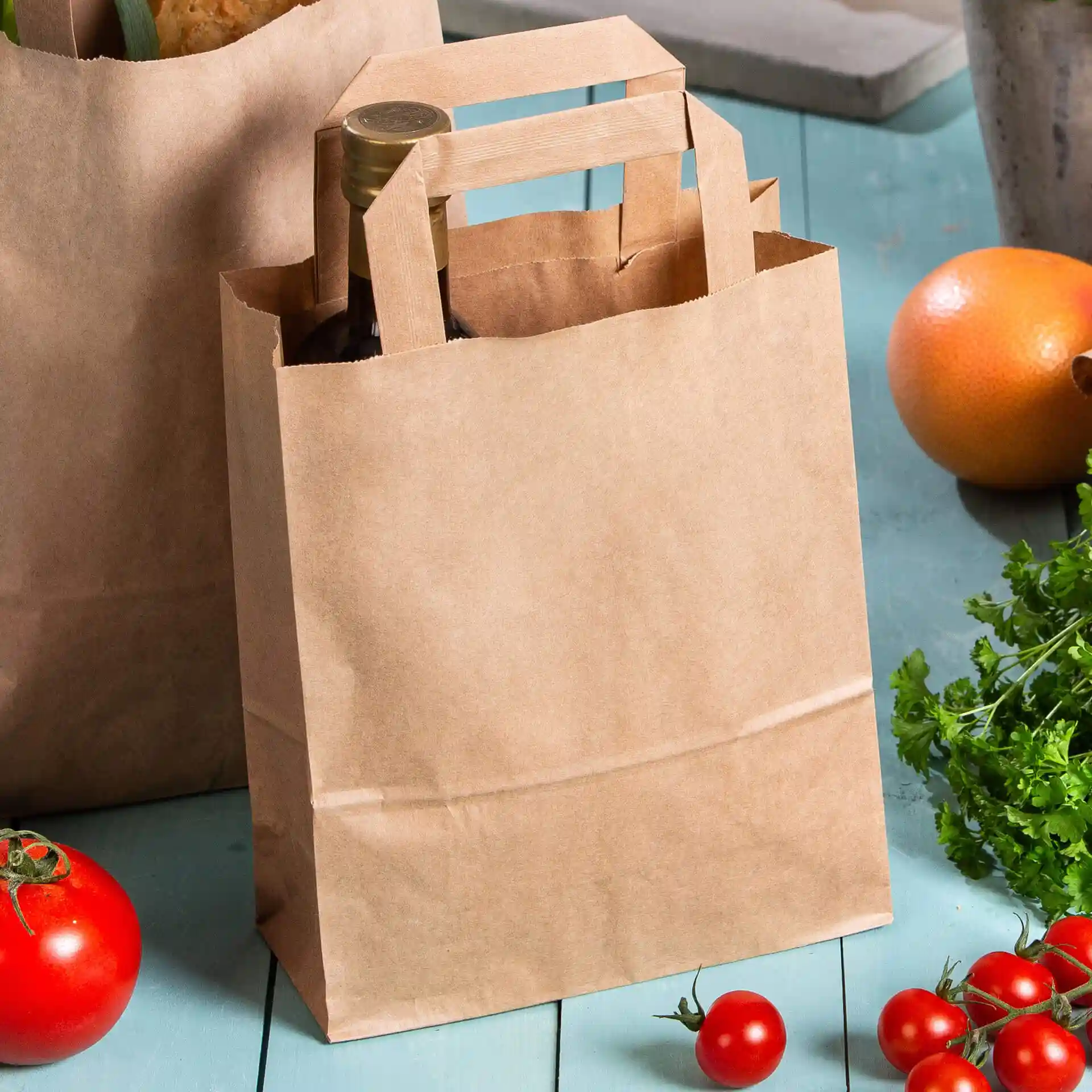 Brązka torba papierowa z uchwytami, zawierająca butelkę, otoczona pomidorami i innymi produktami.
