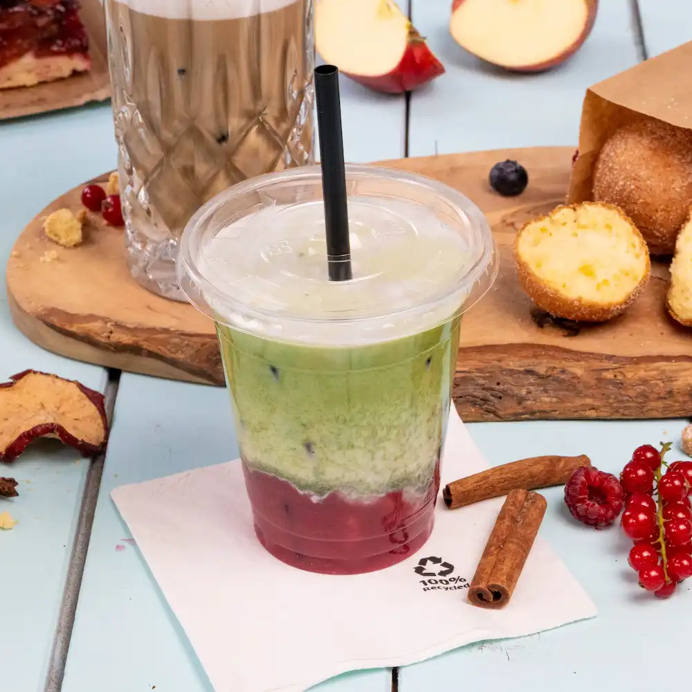 Zimny napój warstwowy w przezroczystym kubku ze słomką, otoczony przekąskami i serwetką na jasnoniebieskiej powierzchni.