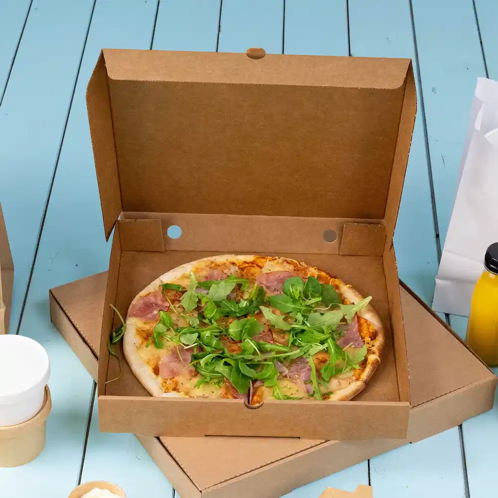 Pizza w kartonowym pudełku z szynką i mieszanką sałat na niebieskiej drewnianej powierzchni.