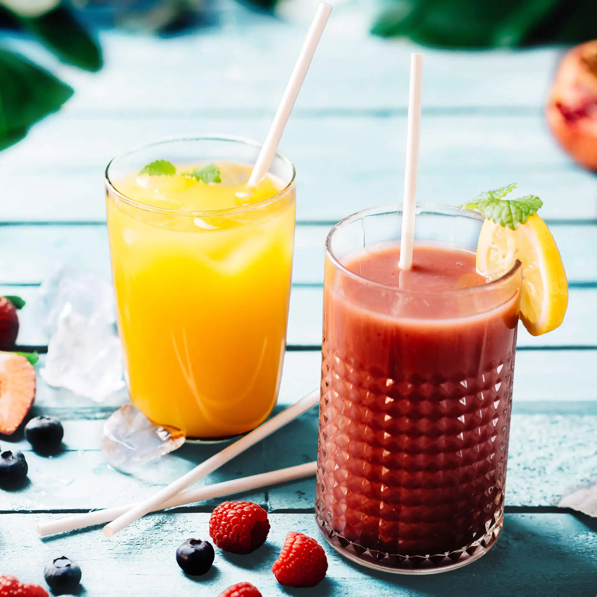 Dwa kieliszki zimnych napojów na drewnianym stole: jeden z żółtym napojem i lodem, drugi z czerwonym napojem udekorowanym cytryną. Białe biodegradowalne słomki i świeże jagody rozsypane wokół.